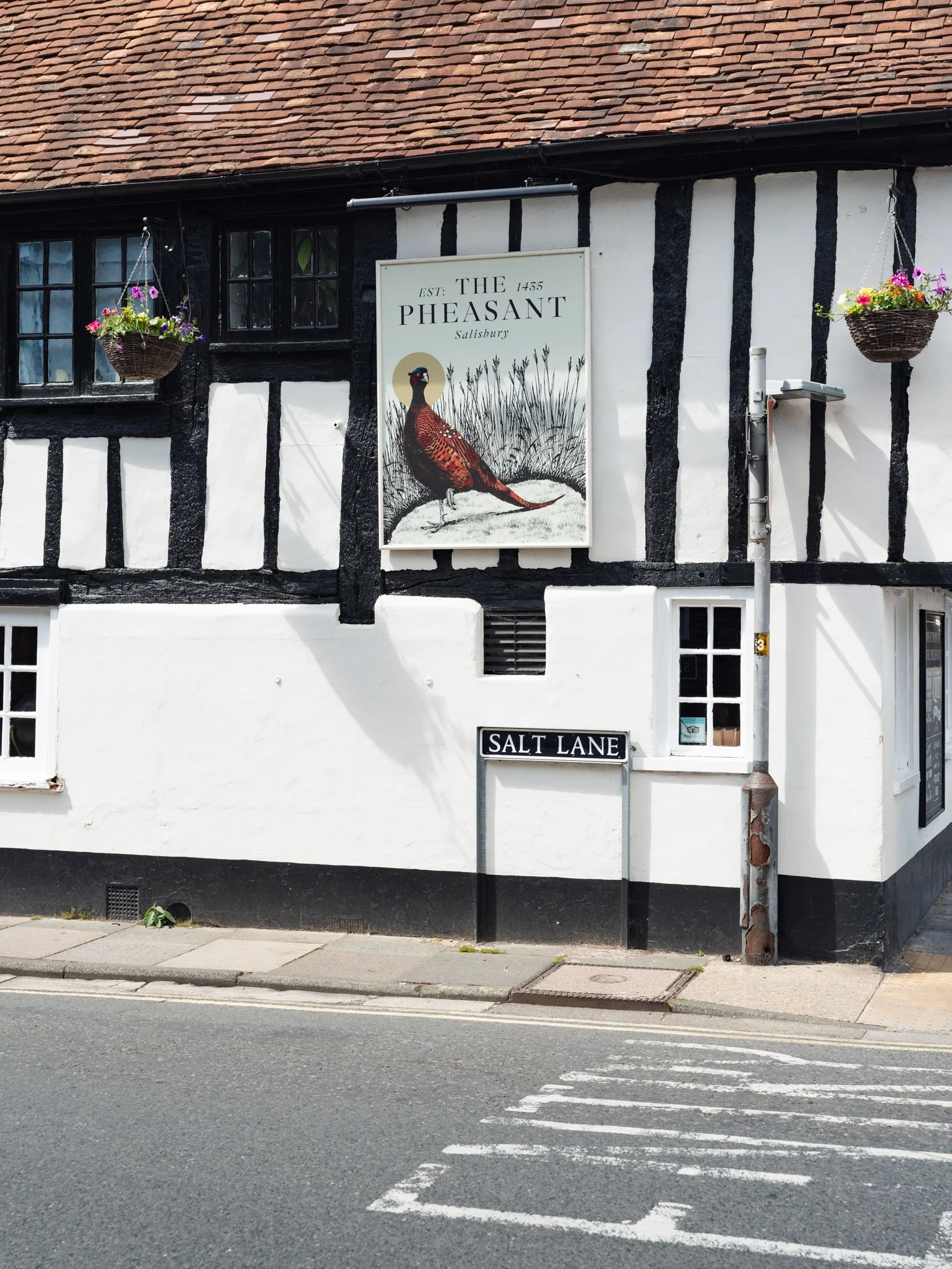 A view of a building with a sign that reads 'The Pheasant Salisbury' and a street sign for Salt Lane. The building has a white exterior with black timber framing, small black window frames, and hanging flower baskets. The street has a pedestrian cros