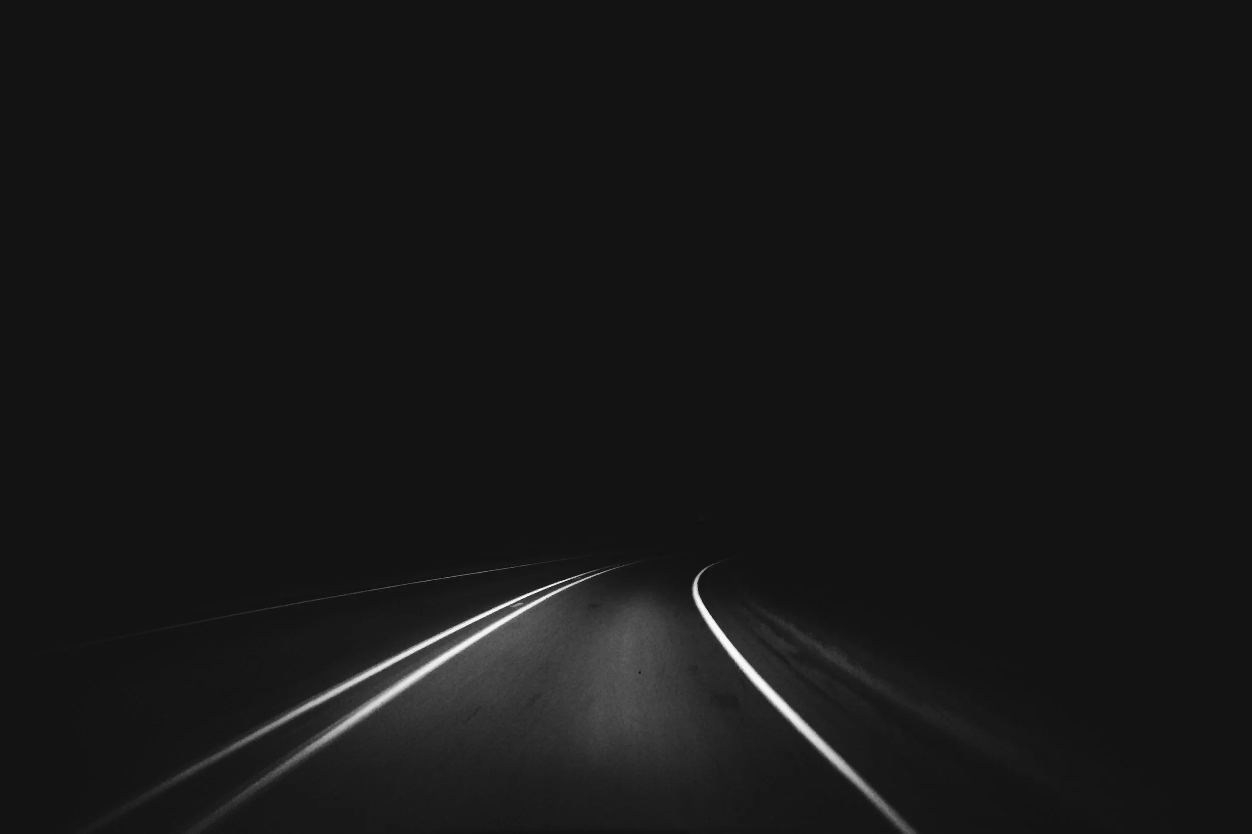 A dark night scene of a road with white lane markings curving into the distance, illuminated by the headlights of a vehicle.
