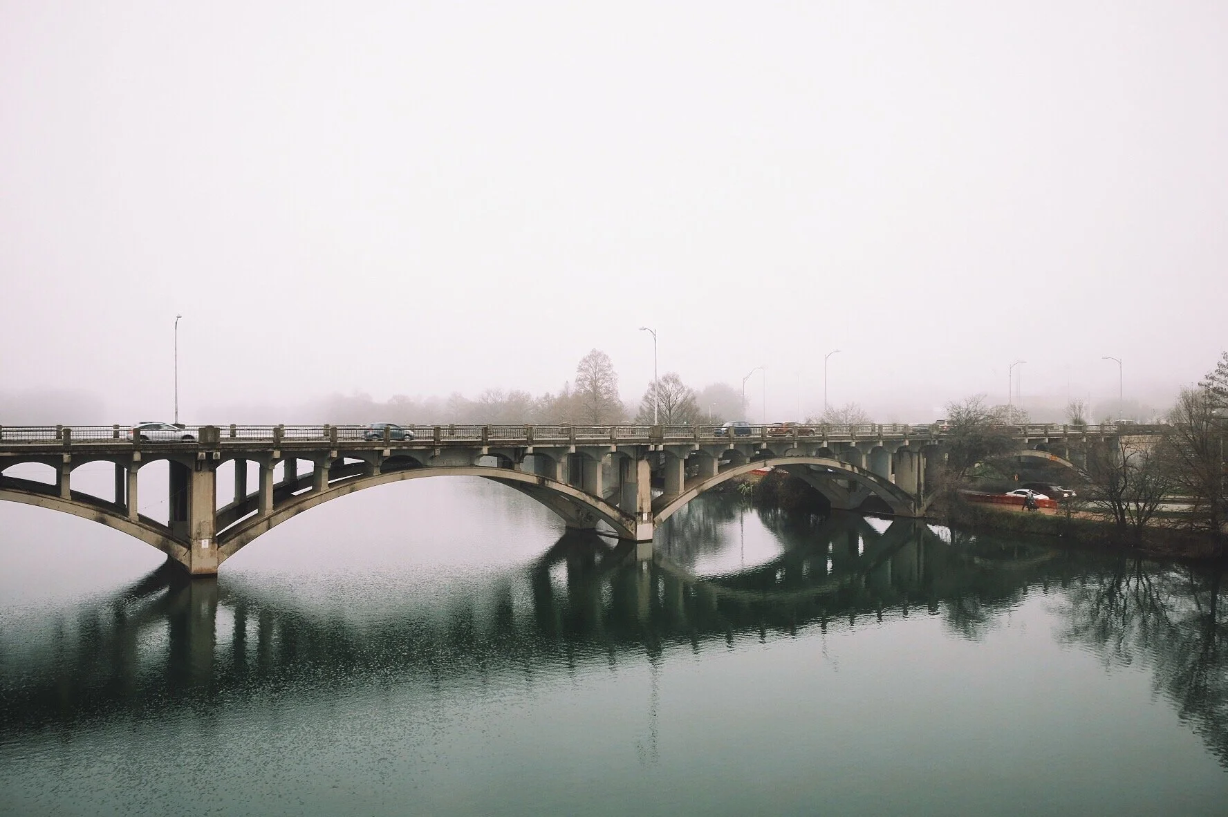 A concrete bridge with multiple arches crossing a body of water, with cars driving on it and trees in the background on a foggy day.