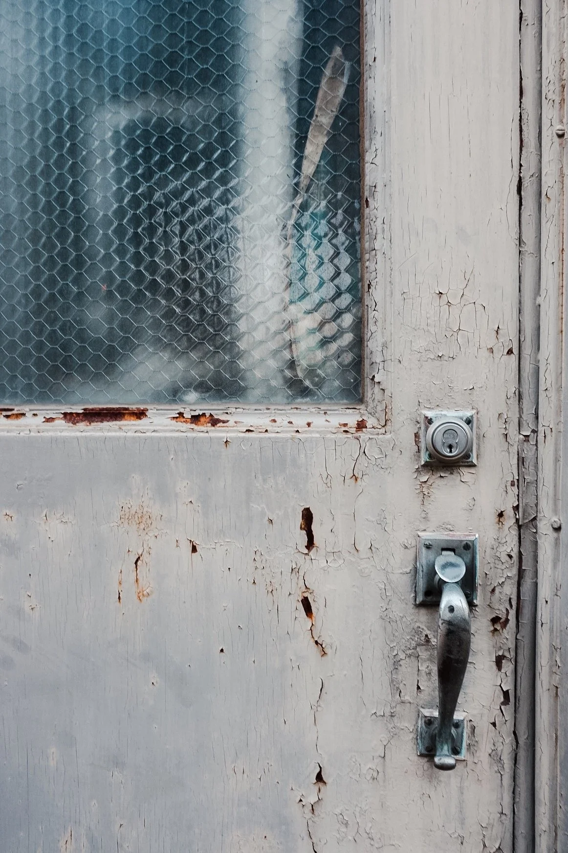 An old, weathered white wooden door with peeling paint, a frosted glass window with a honeycomb pattern, a rusted lock, and a metal handle.