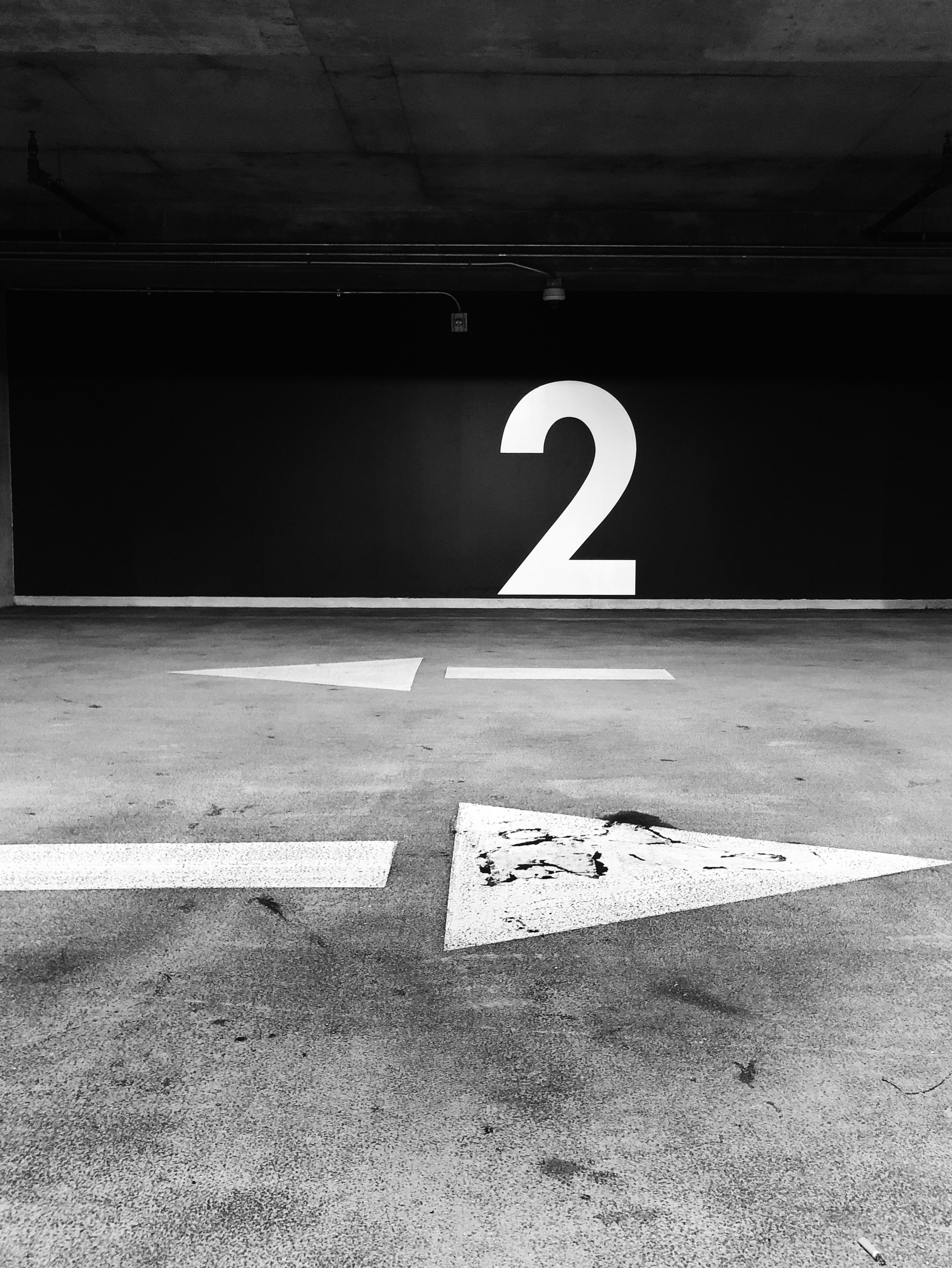 Black and white photo of a parking garage with a large white number '2' on a wall and a faded arrow painted on the ground.