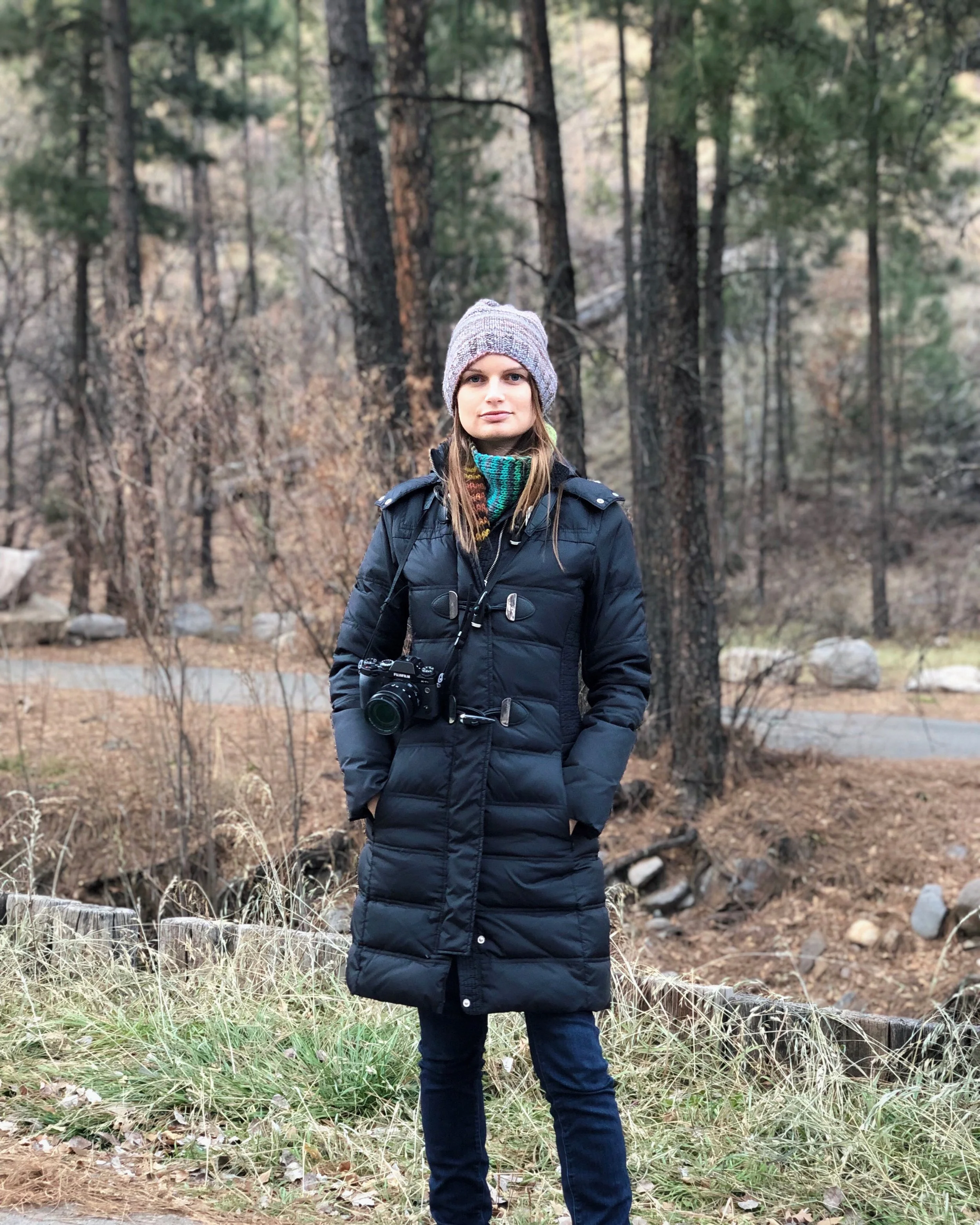 Woman in black winter coat, knit hat, and jeans standing outdoors in a forested area with tall trees and dry grass.