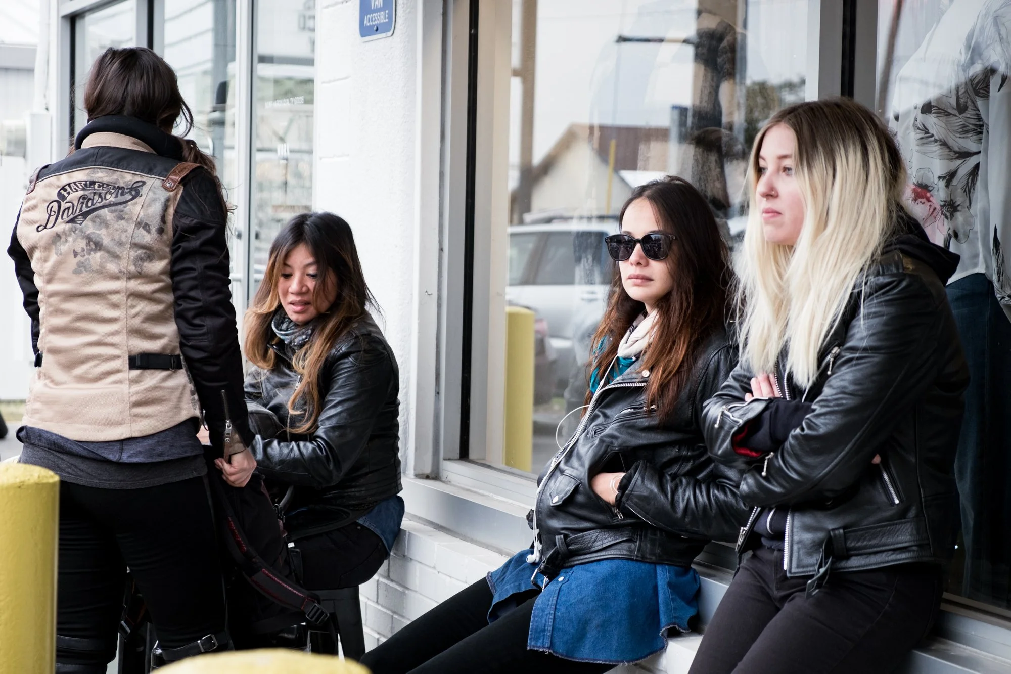 Four young women waiting outside a building; some sitting on a bench and others standing, dressed in casual clothing and leather jackets.
