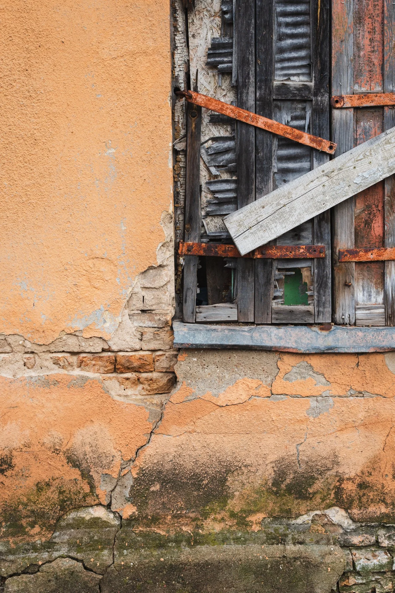 The image shows an old, weathered wall with peeling peeling orange paint, cracks, and exposed bricks. A section of the wall features a boarded-up window with old, gray, and rusted wooden and metal shutters.