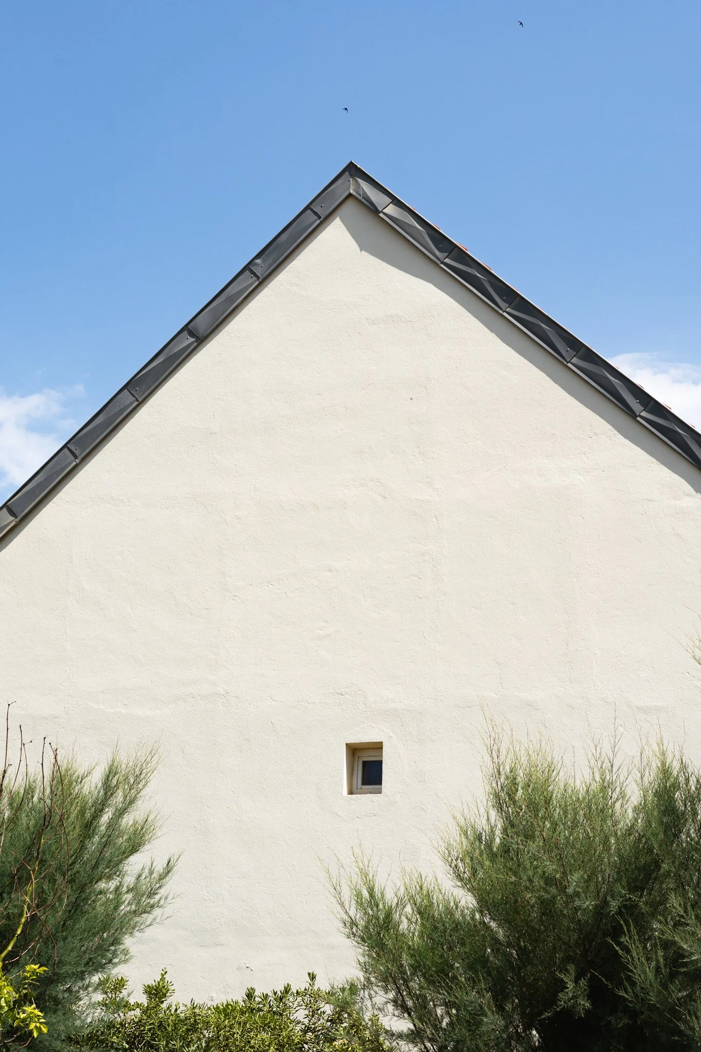 A white building with a triangular roof and small window, surrounded by green shrubbery, under a clear blue sky with birds.