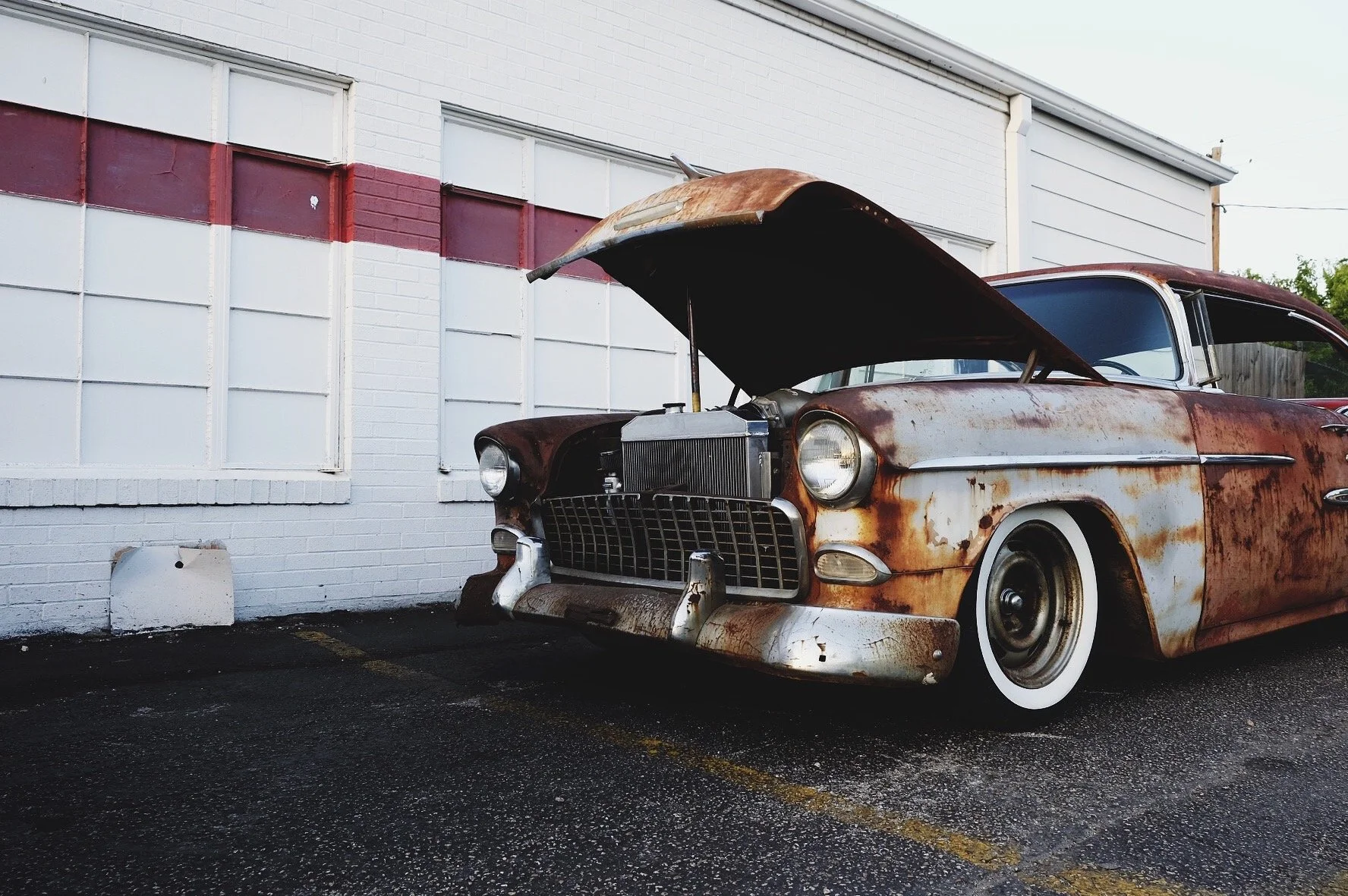 Rusty, abandoned vintage car with open hood parked on wet asphalt in front of a white brick building with boarded windows.