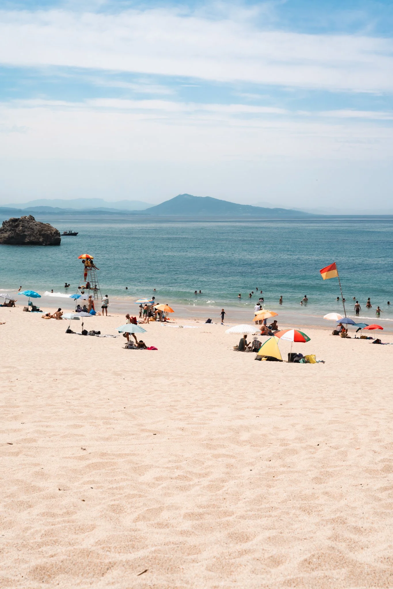 A crowded beach with umbrellas, swimmers, a lifeguard tower, and a German flag near the shoreline. Mountains are visible in the background under a partly cloudy sky.
