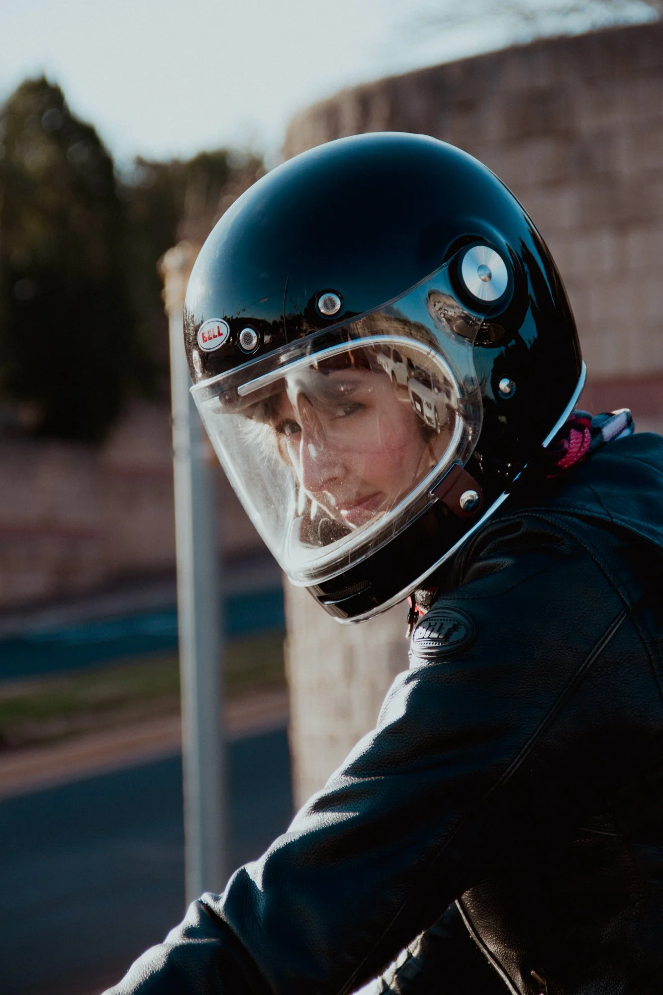 A person wearing a black motorcycle helmet and leather jacket, leaning against a brick wall outdoors, with blurred trees and sky in the background.