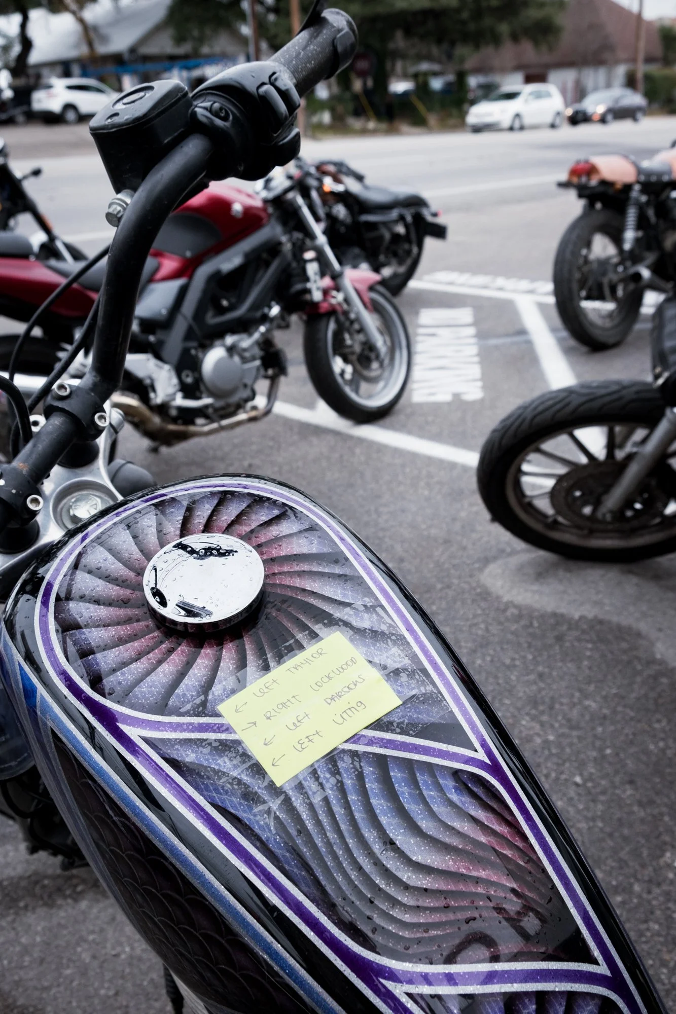 Close-up of a motorcycle fuel tank with a purple and black custom paint job, rain droplets, and a sticky note with directions. In the background, there are several other motorcycles and cars parked on the street.