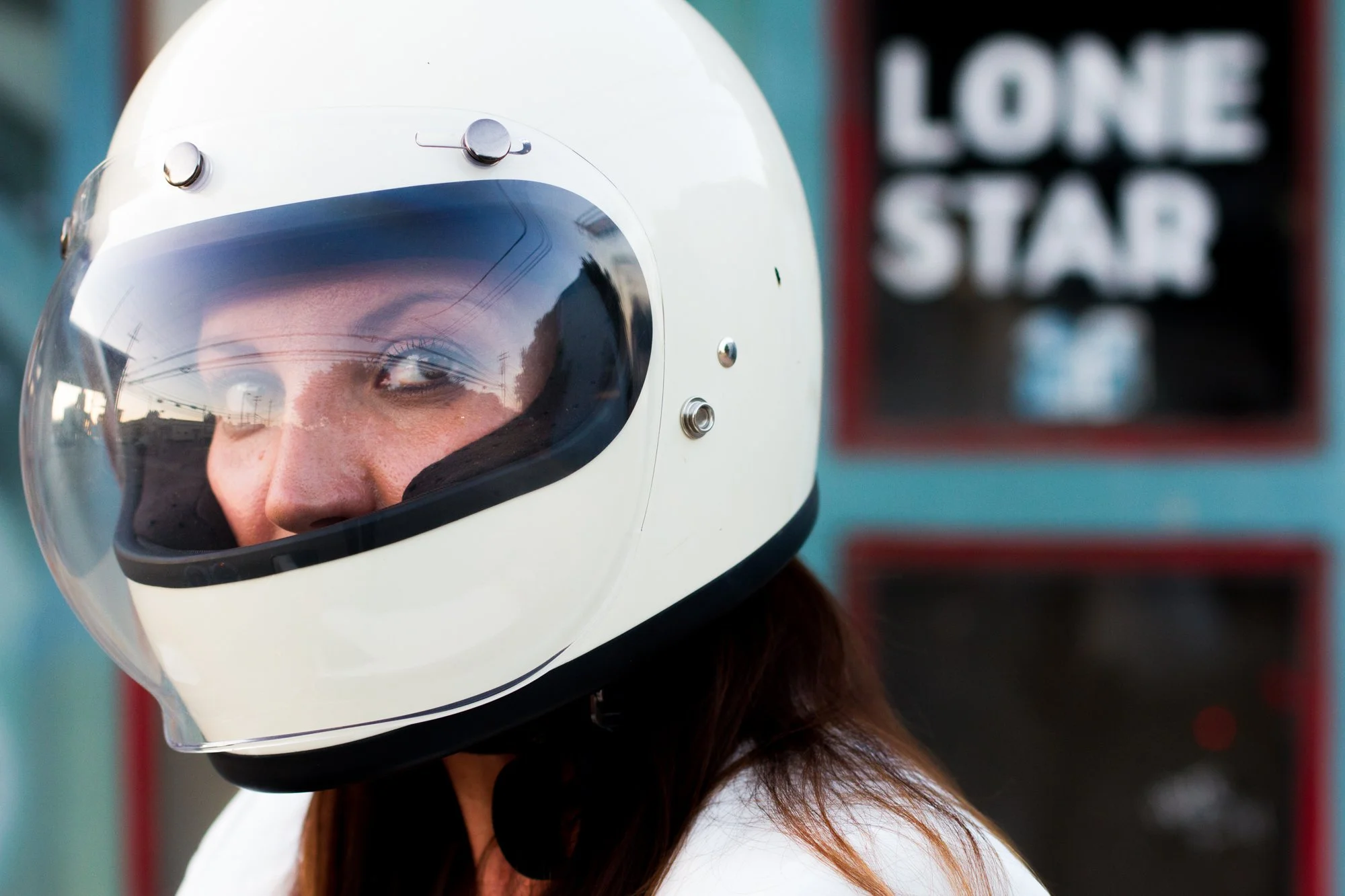 A woman wearing a white motorcycle helmet with a clear visor, looking to her left, with a blurred sign reading 'LONE STAR' in the background.