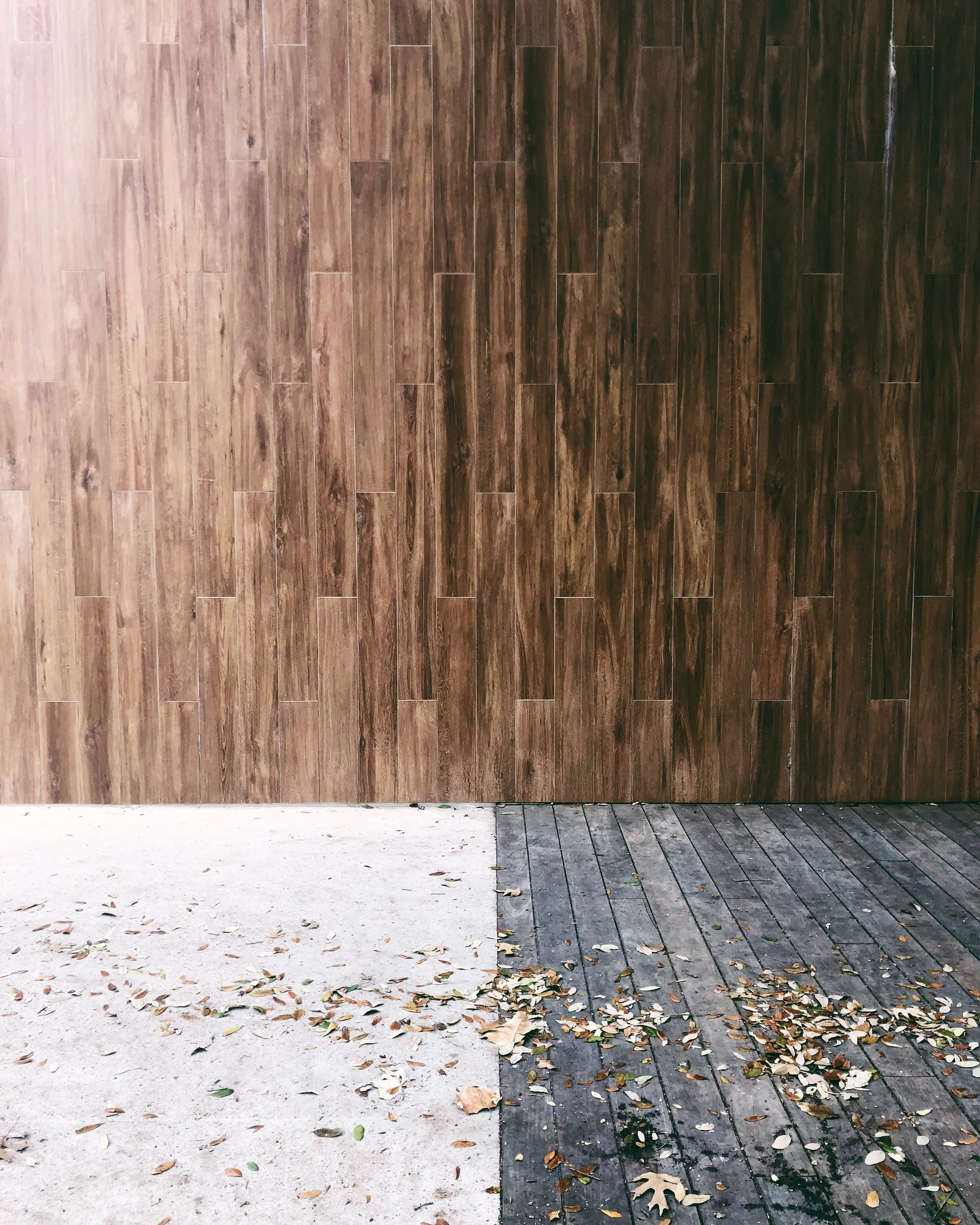A large wooden wall with a sidewalk at its base. The sidewalk is partially covered with fallen leaves. The wall is made of long, narrow wooden panels with a dark brown finish.