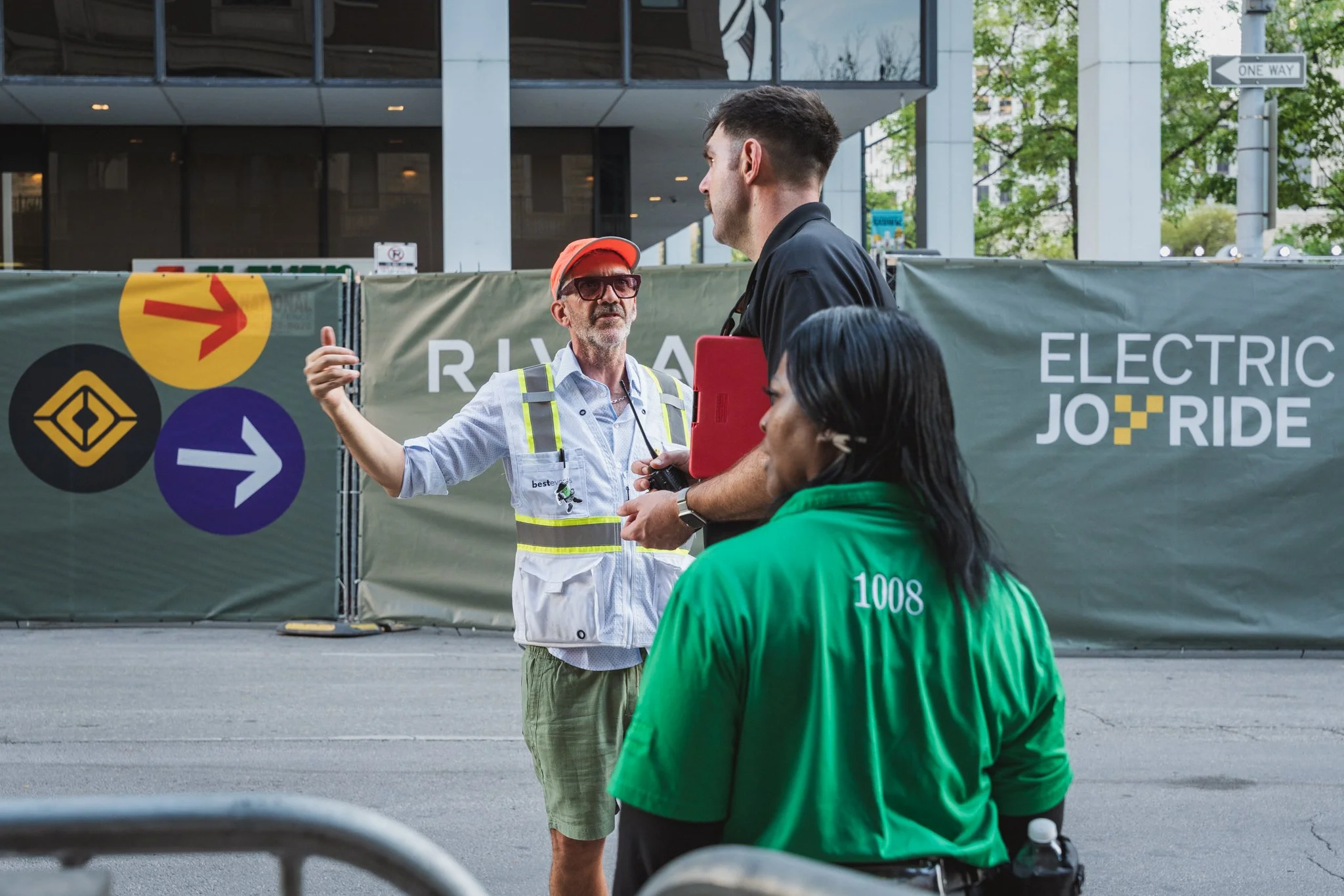 A man in a black shirt is speaking to a worker at an Electric JF Ride event, with a woman in a green jacket nearby. The worker is wearing a staff badge, safety vest, and a red cap with glasses, outside a building with glass windows and a colorful ban