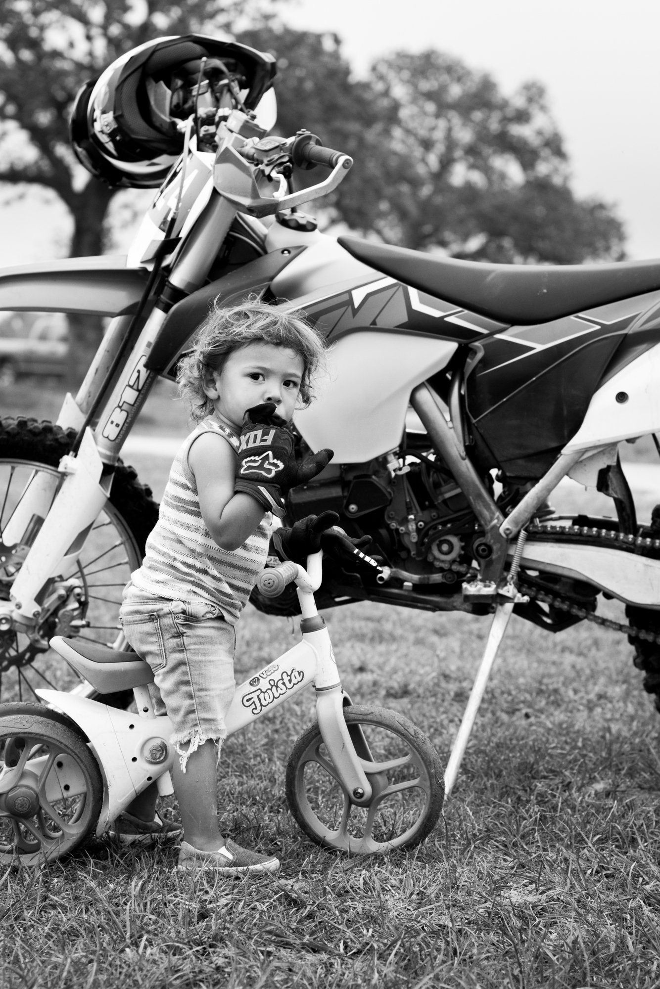 A young girl on a small balance bike, standing next to a dirt bike, touching her lips with her finger, outdoors with trees in the background.
