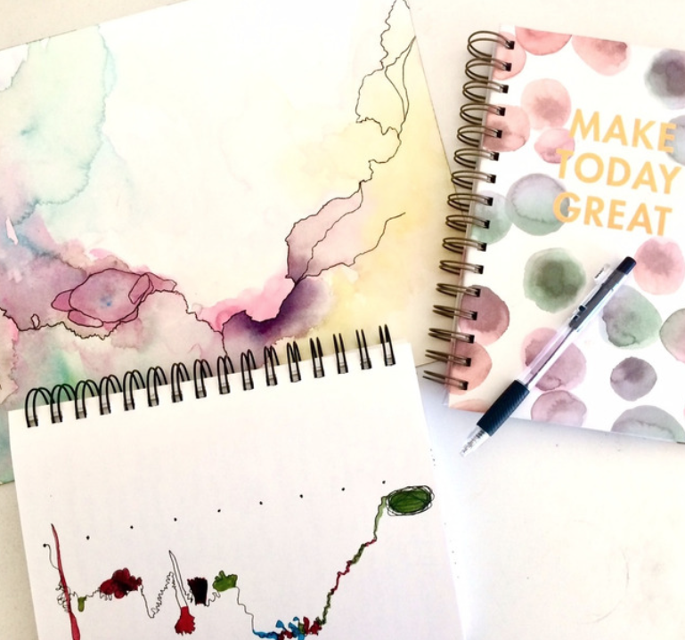 Top-down view of three open journals on a tabletop, featuring colorful watercolor splashes, expressive line work, and a pen resting on the middle journal. One cover reads "MAKE TODAY GREAT," adding an uplifting touch to the creative scene.
