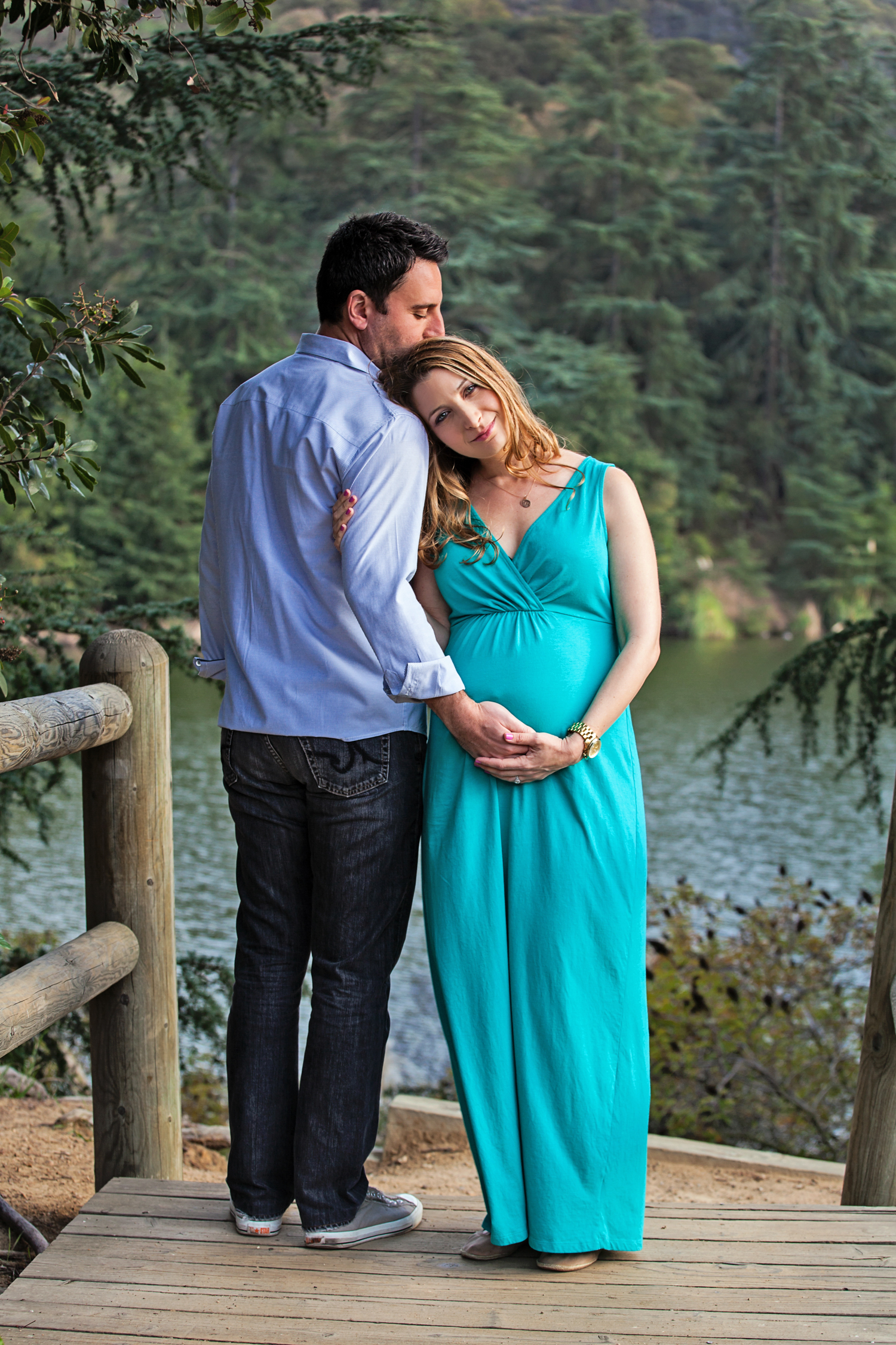 happy pregnant couple wearing shades of blue in front of lake 