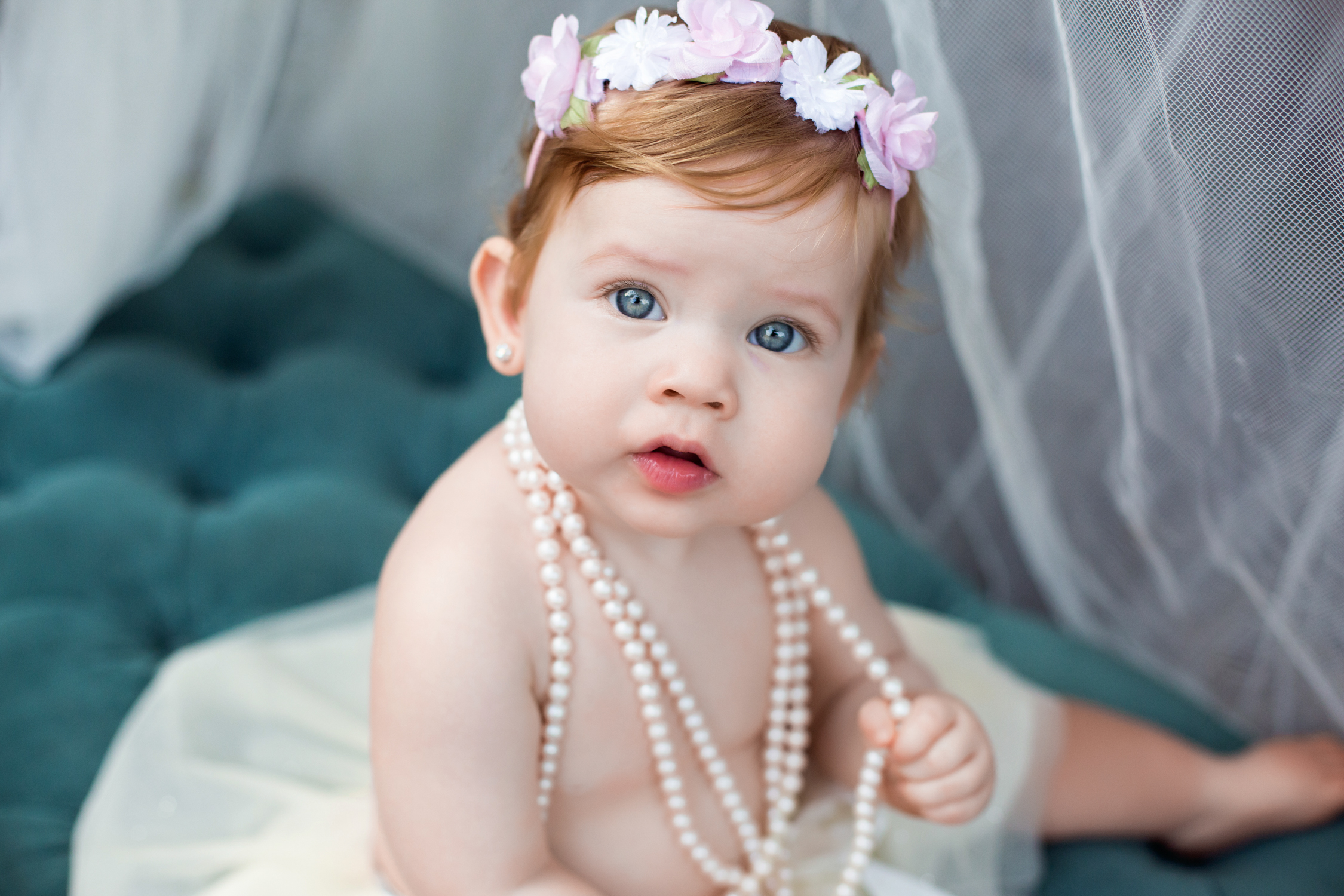 red haired baby girl in a tutu and flower headband holds the pearl necklace she is wearing