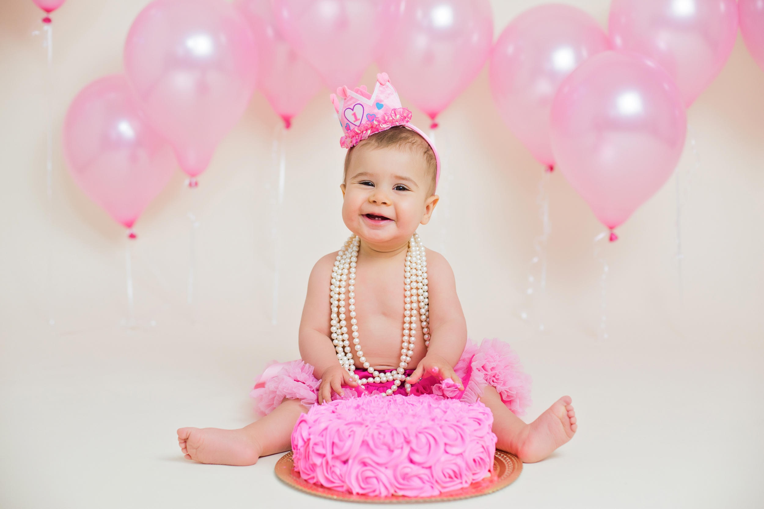 birthday baby girl sitting behind a hot pink cake, with pink balloons behind 