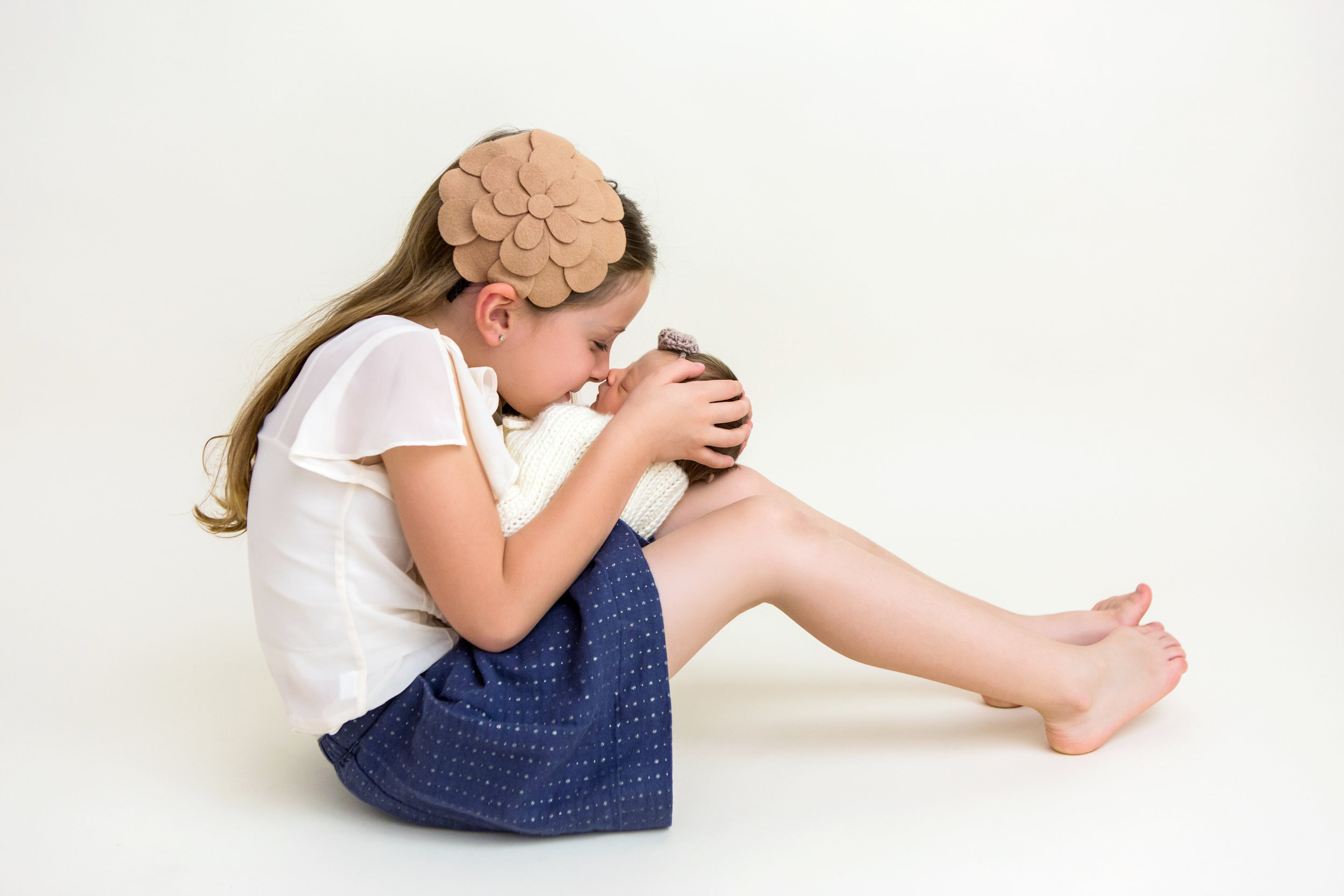 big sister with a brown flower headband snuggling close with her baby sister, wrapped in white