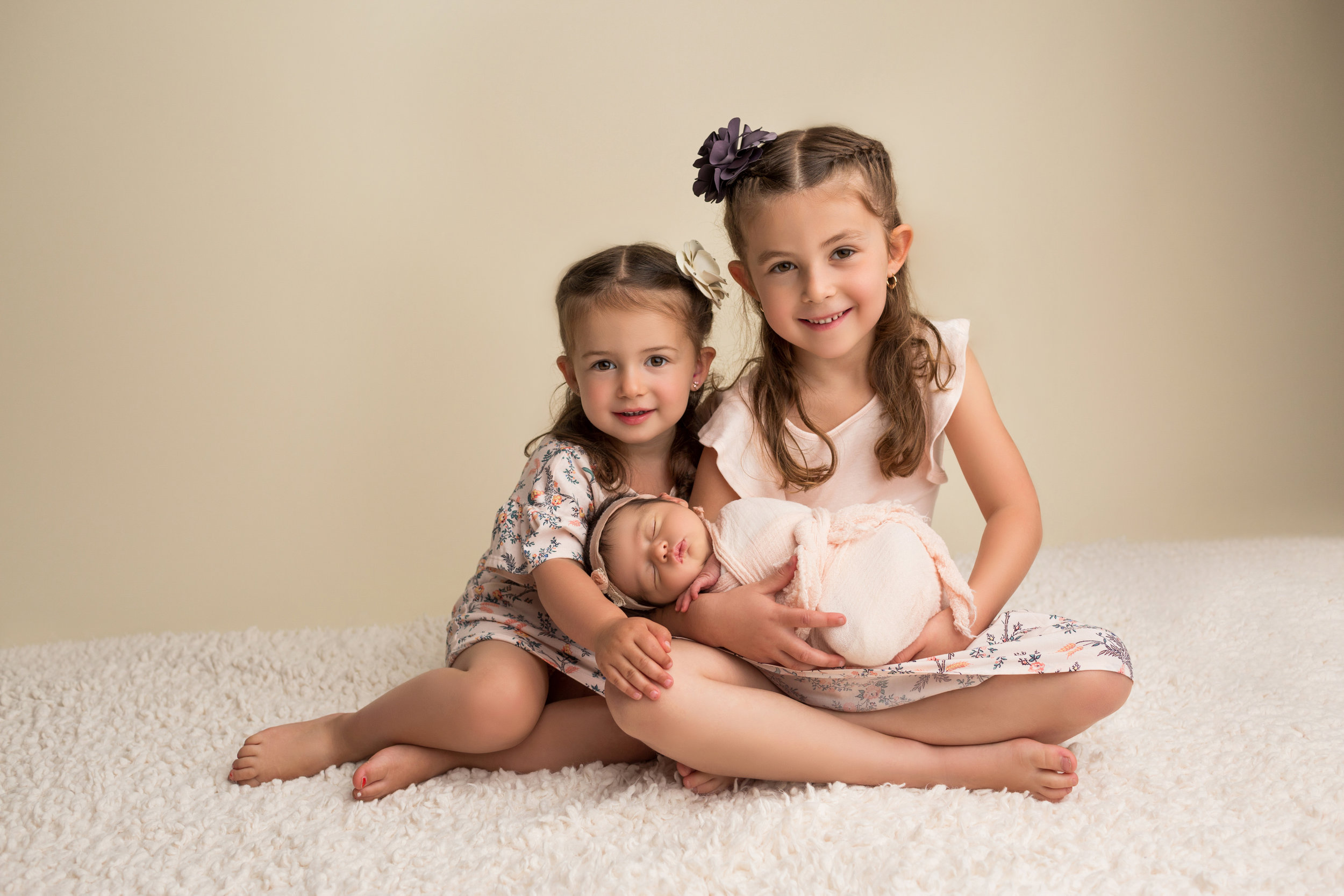 two sisters lovingly hold their newborn sister while sitting on a white rug