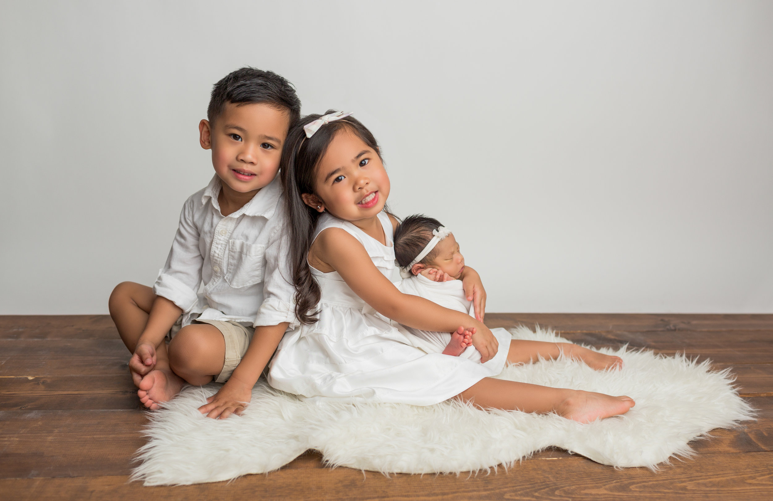 brother and sister with their newborn sister on a white rug