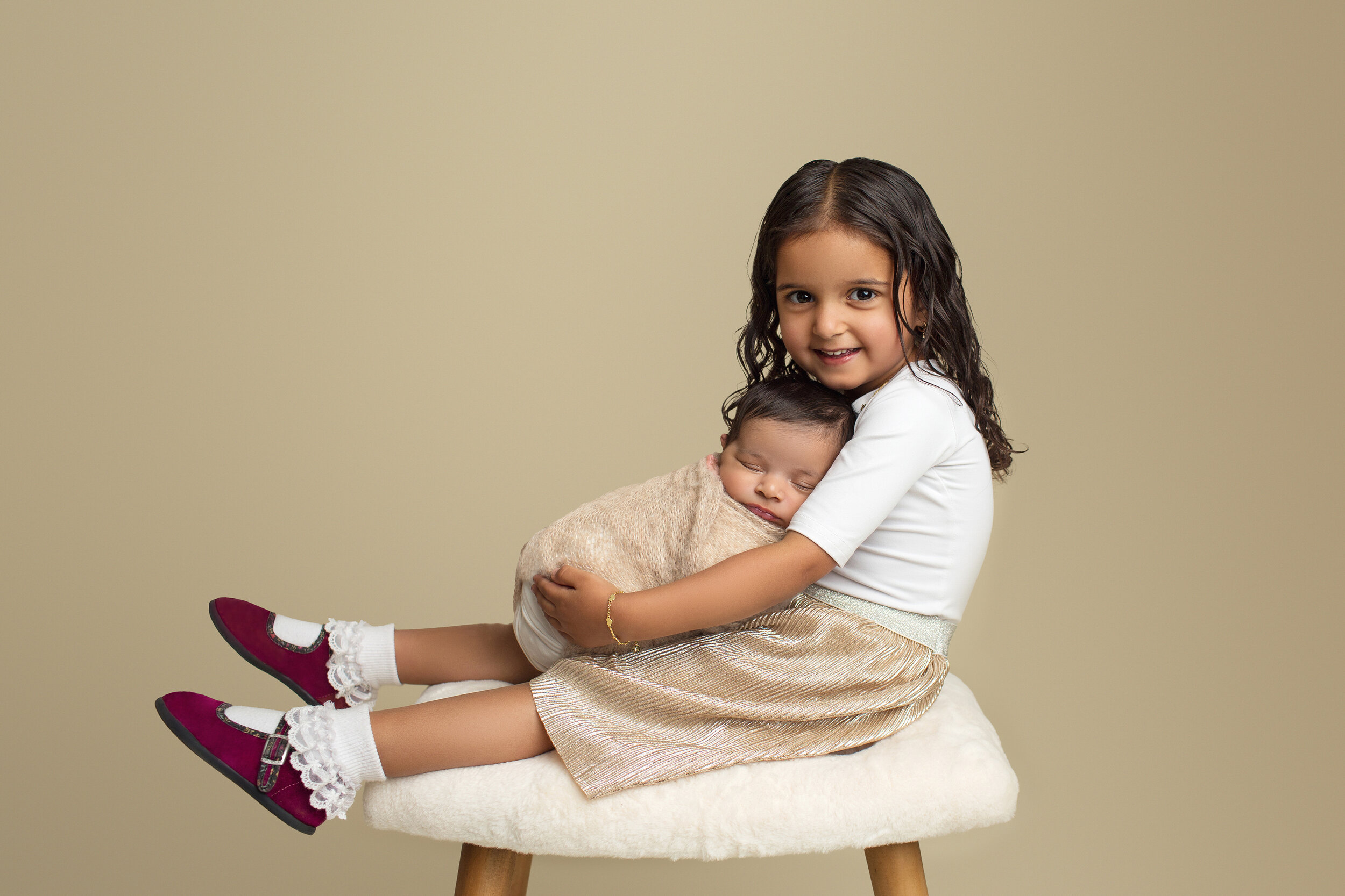 big sister holds her newborn sister on her lap while sitting on a white bench