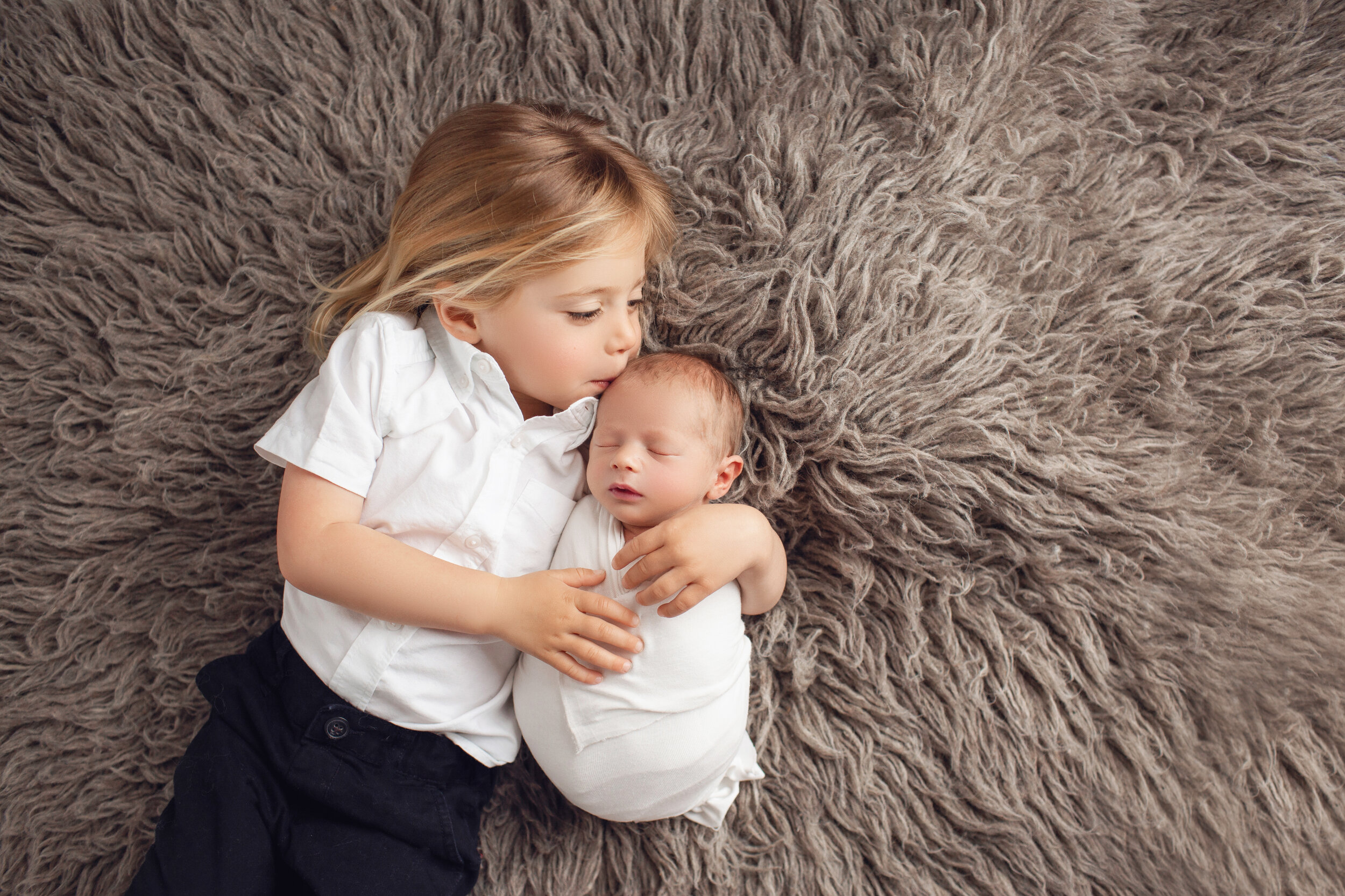 while lying on a brown rug, big brother kisses his baby brother sweetly