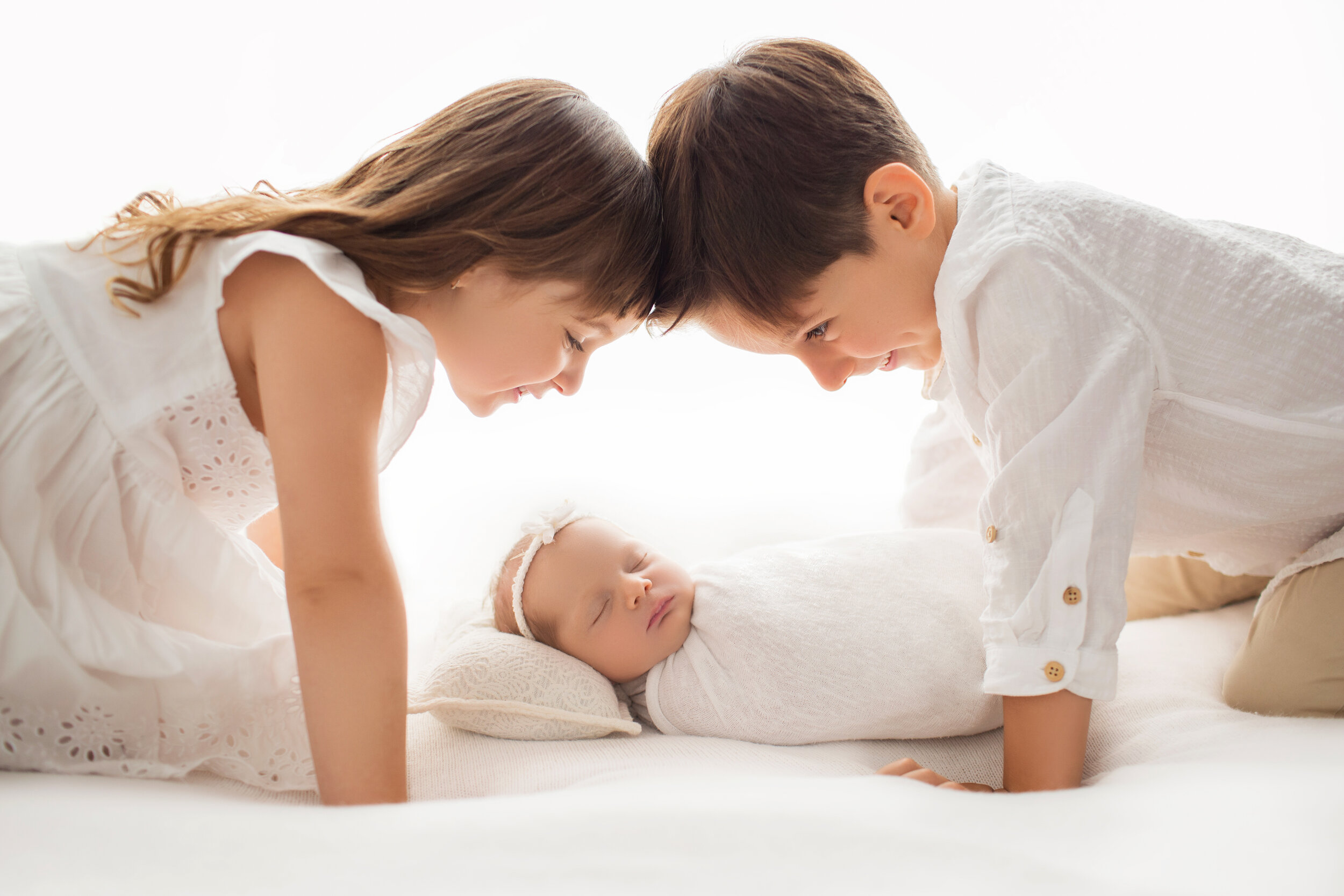 older brother and sister look lovingly down at their newborn sister