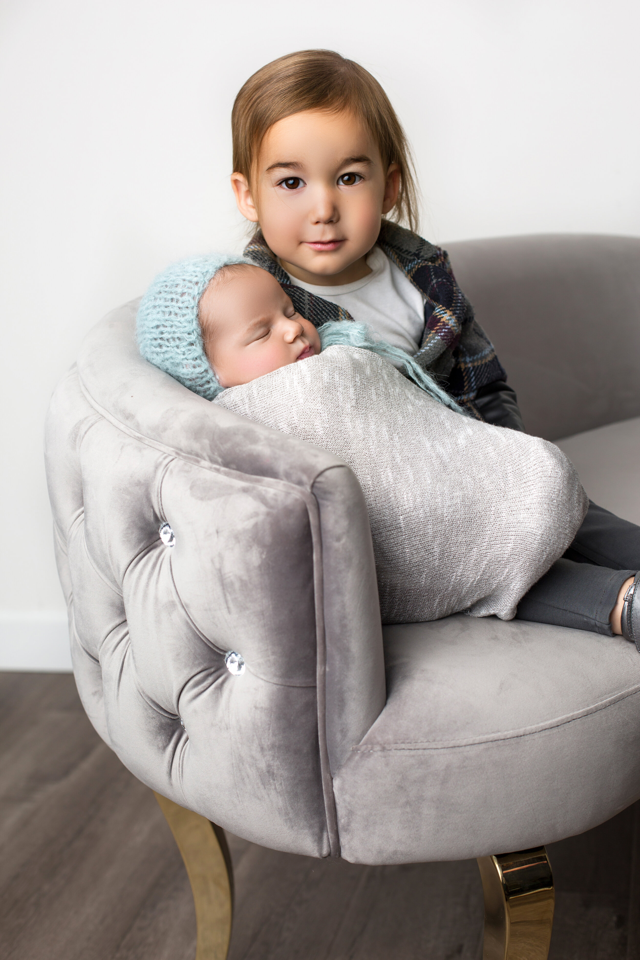 brother sits next to his newborn brother, who is propped against the back of the chair