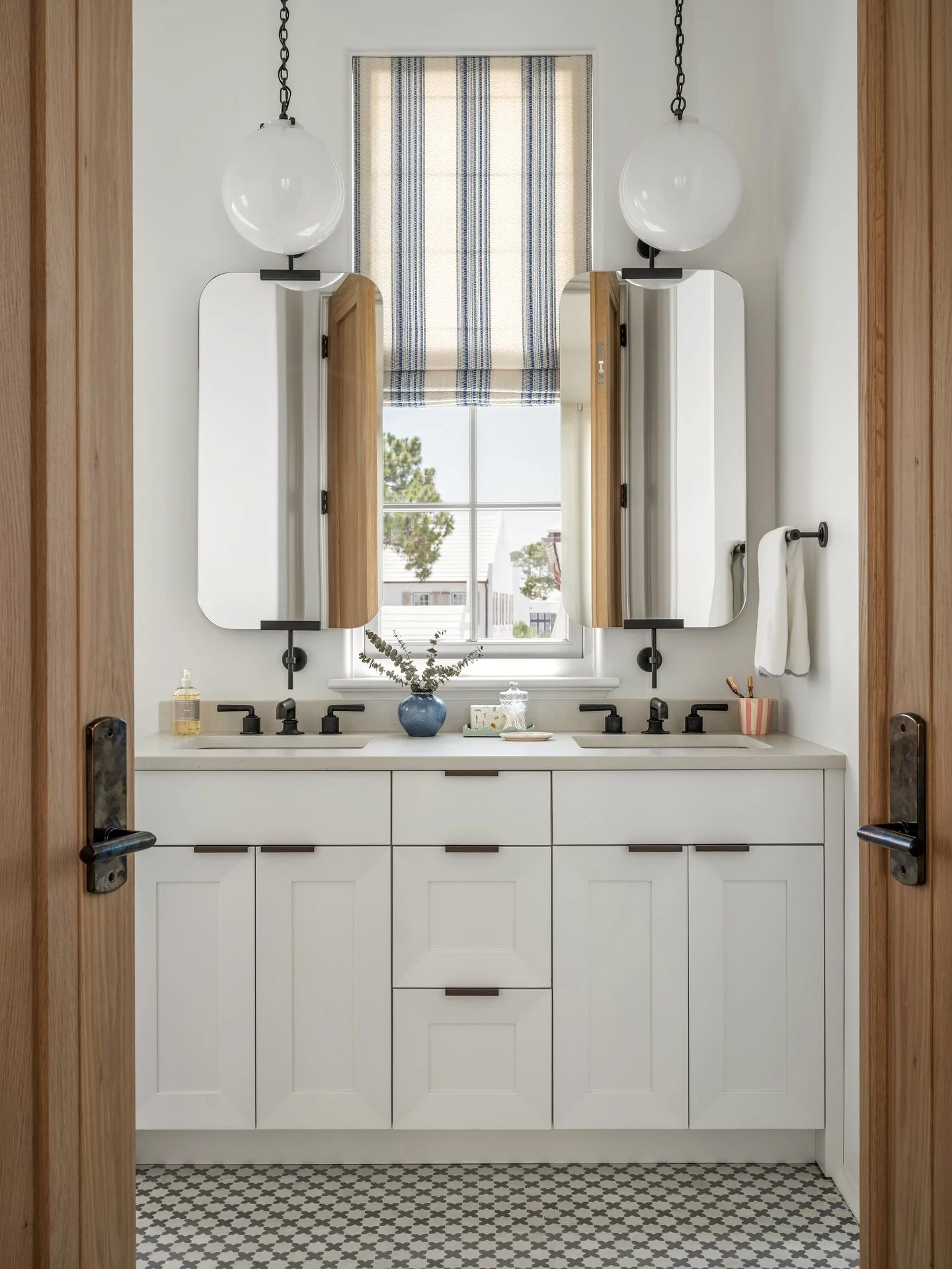 ✨🌊 Patterned tile floors bring character underfoot, while custom inset cabinetry provides a seamless, tailored look. Soft Roman shades filter natural light, adding warmth against the clean lines of matte black fixtures. Globe pendants frame the mirr