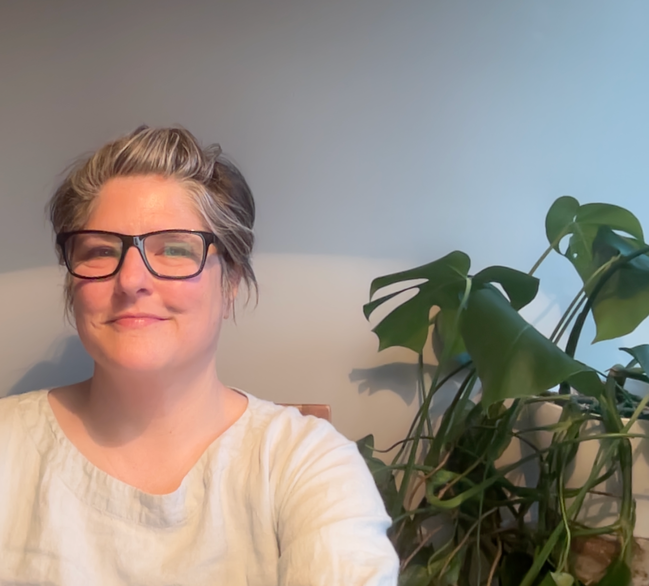 White woman wearing glasses, smiling and sitting next to a plant