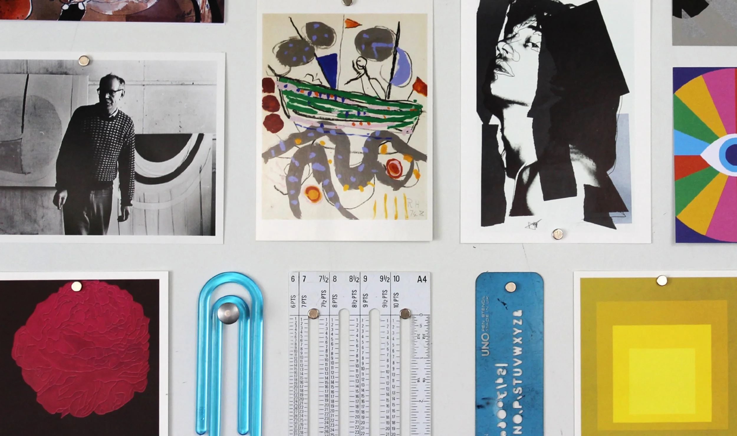 A wall display with various art prints, a blue plastic ruler, a color wheel, a black-and-white photograph of a man, and a red textured circular art piece.