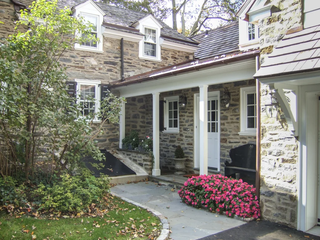 Stone house with small front porch, white trim, pink flowers, bushes, and a black bench.