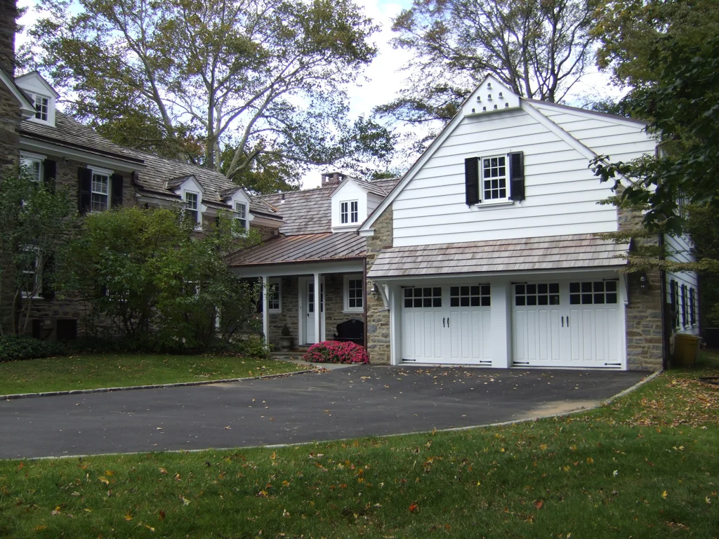 A house with a stone and white exterior, black shutters, a two-car garage, and a driveway, surrounded by trees and grass.