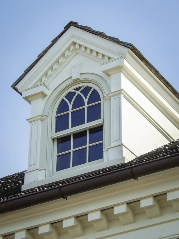 Close-up of a house's decorative attic window with moldings and trim, part of a white house with a dark roof.