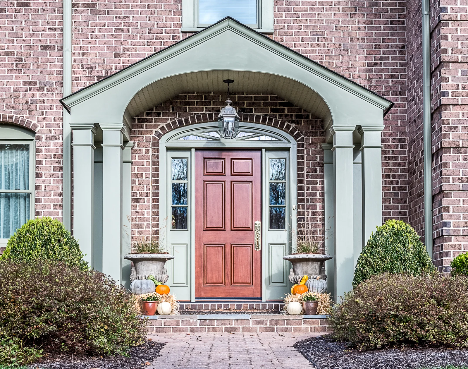 Front entrance of a brick house with a red door, decorative planters, pumpkins, and a lantern hanging above the door.