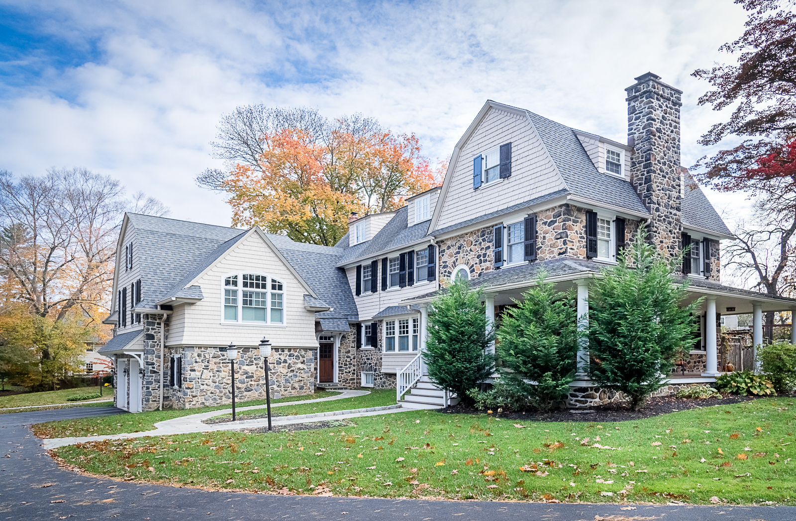 Large house with stone and siding exterior, surrounded by trees with fall foliage, green lawn, and a curved driveway with three black lamp posts.