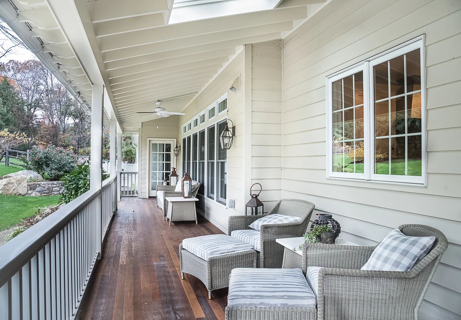 Covered porch with outdoor furniture including wicker chairs, a small table, pillows, lanterns, and a ceiling fan, overlooking a garden with rocks and trees.