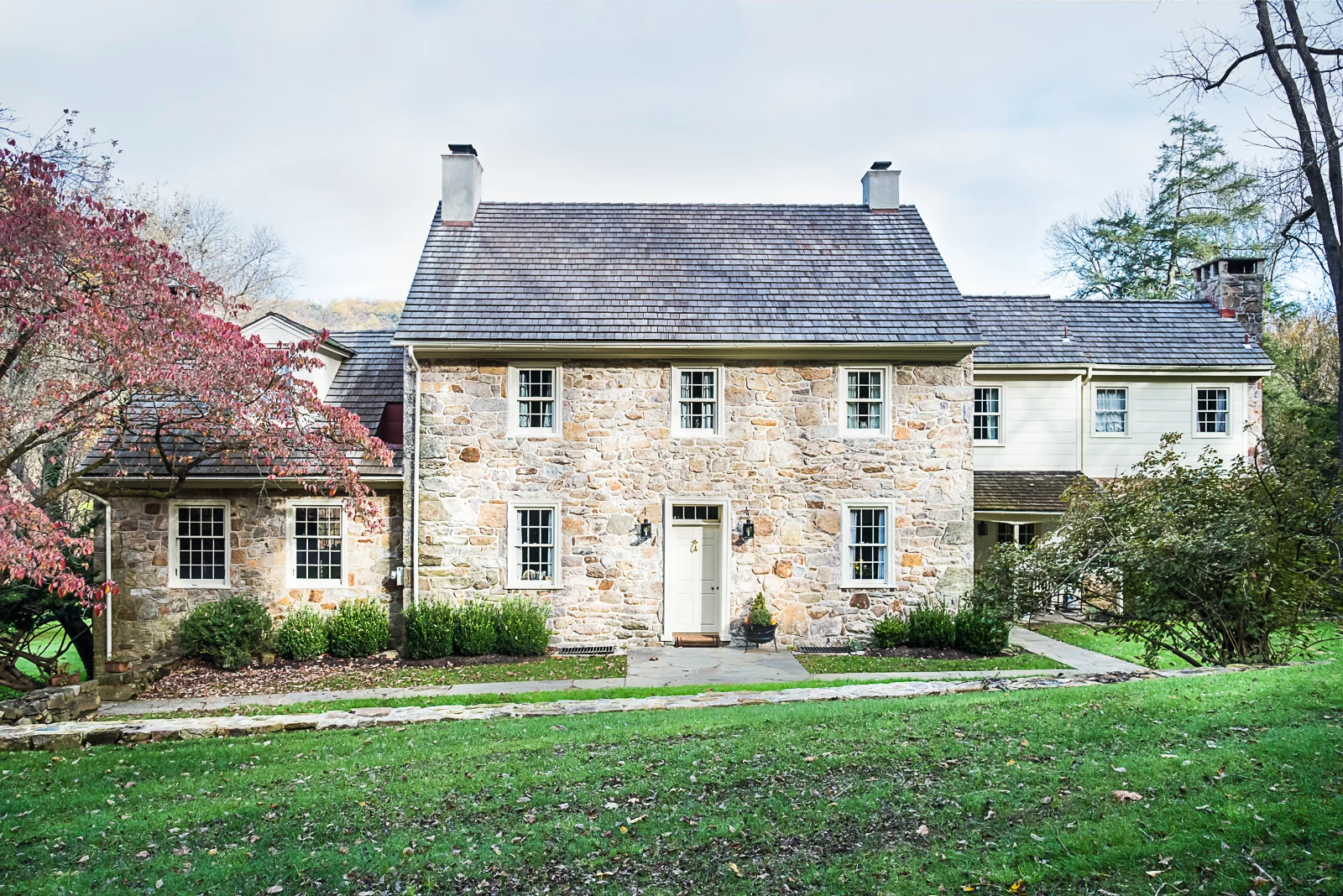 A stone house with a front door, surrounded by grass and trees, some with pink and green leaves, under an overcast sky.