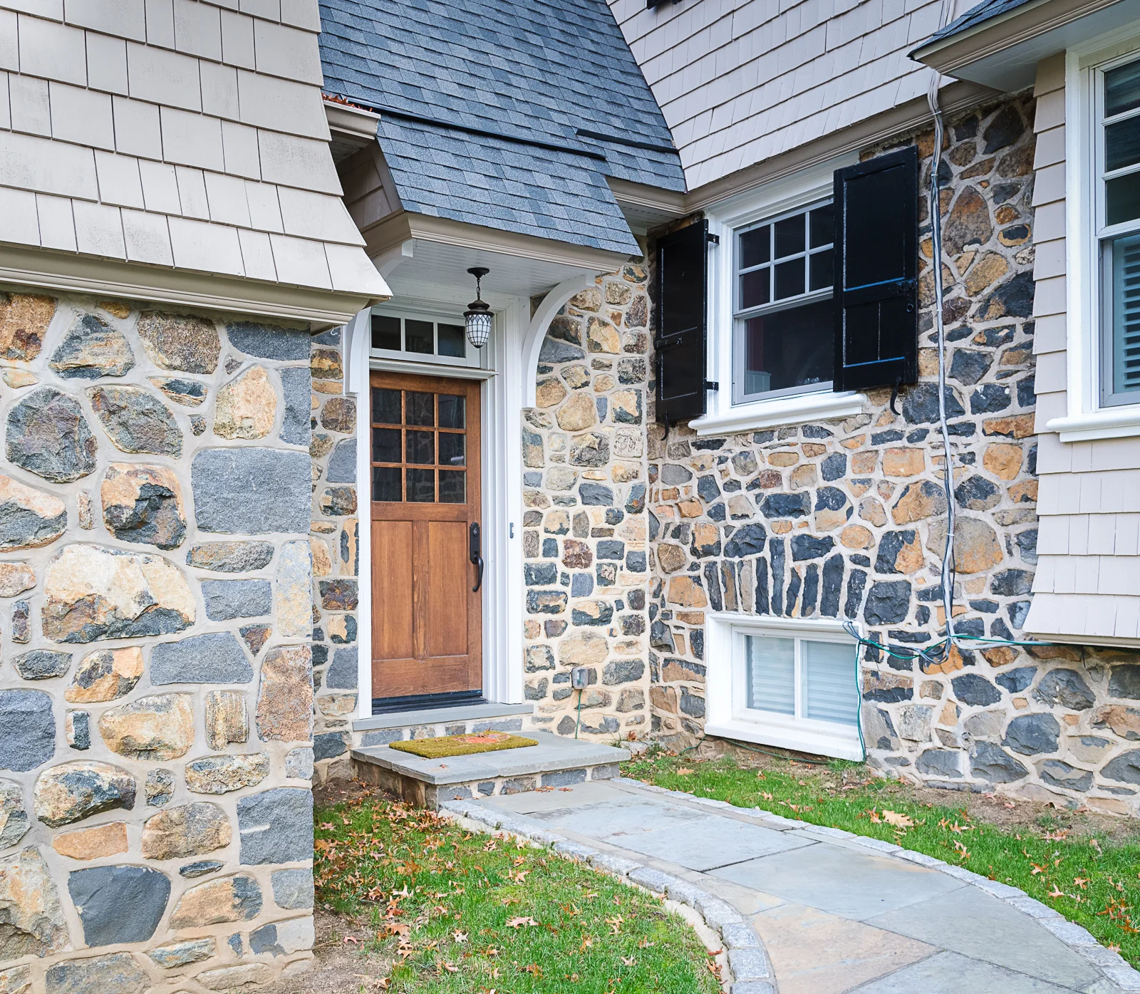 Exterior view of a house with stone and siding walls, a wooden front door with glass panes, black window shutters, a small patio, and a stone pathway.