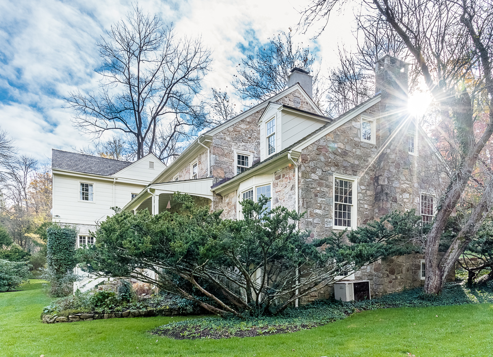 A large stone house with white trim, surrounded by green grass and shrubs, with leafless trees and a partly cloudy sky in the background sunlight shining from the right