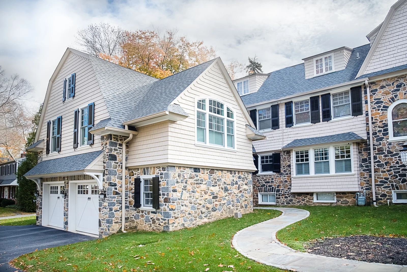 A large multi-story house with stone and white siding exterior, multiple windows with black shutters, and a driveway leading to a double garage. A curved concrete pathway runs through the front lawn.