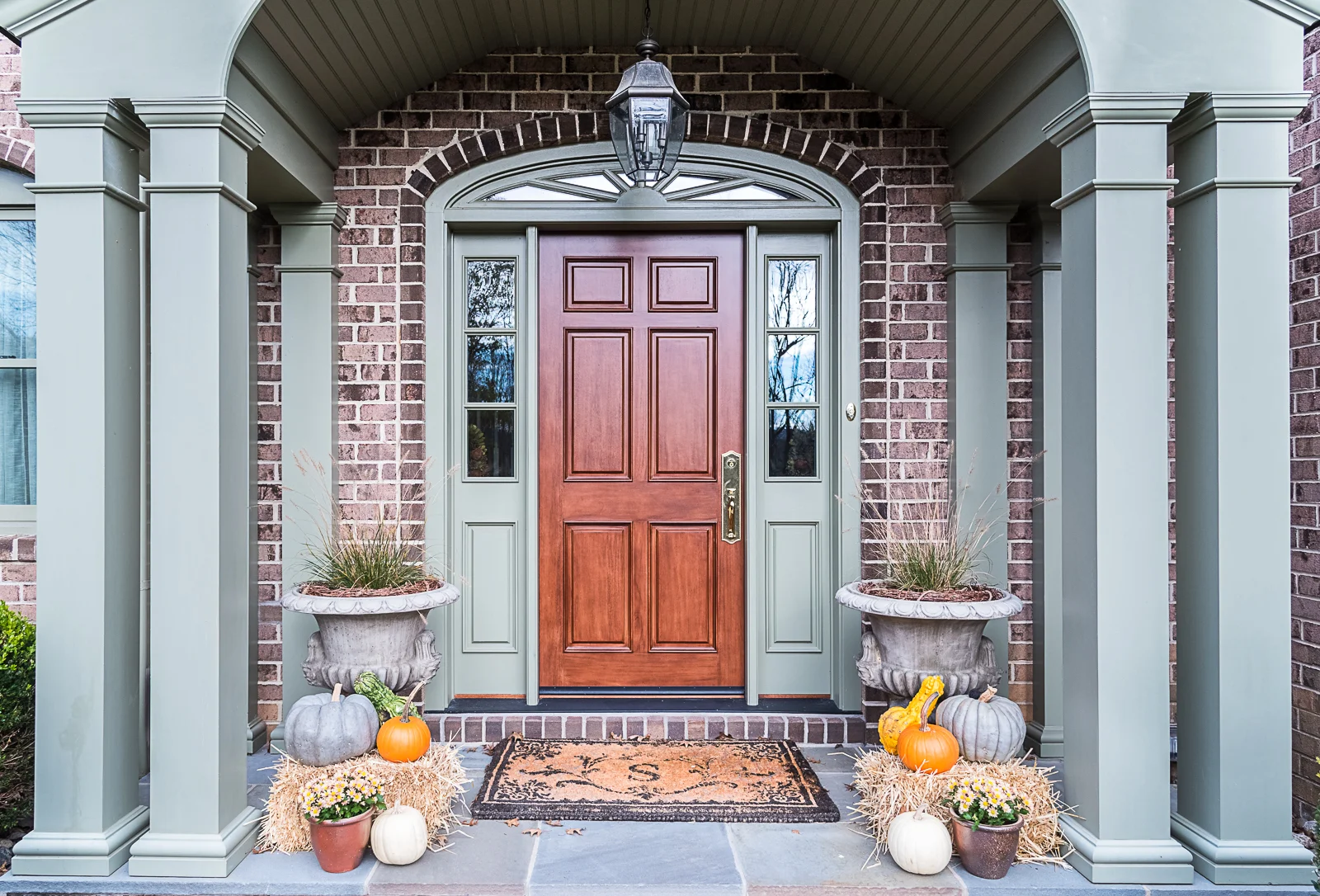 Decorative fall-themed front porch with pumpkins, potted plants, and hay bales, featuring a wooden front door, brick exterior, and columns.