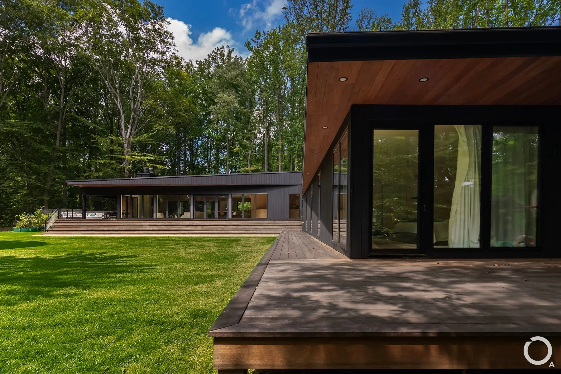 Contemporary home with a broad wood deck, floor-to-ceiling windows, and warm wood soffits overlooking a landscaped lawn and surrounding forest.  Photo Credit: Linda McManus Photography