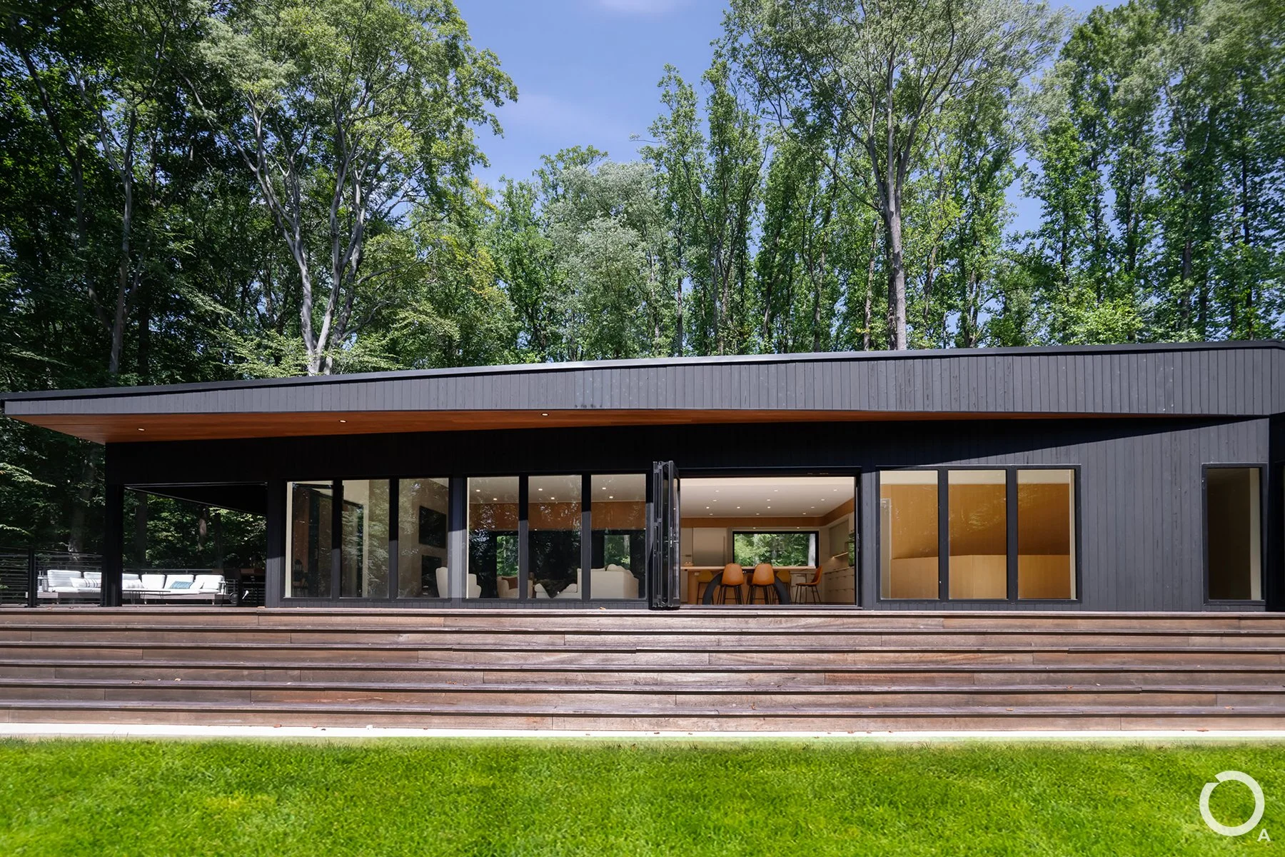 Rear elevation of a modern custom home featuring dark vertical siding, folding glass doors, expansive wood decking, and a deep roof overhang. Photo Credit: Linda McManus Photography