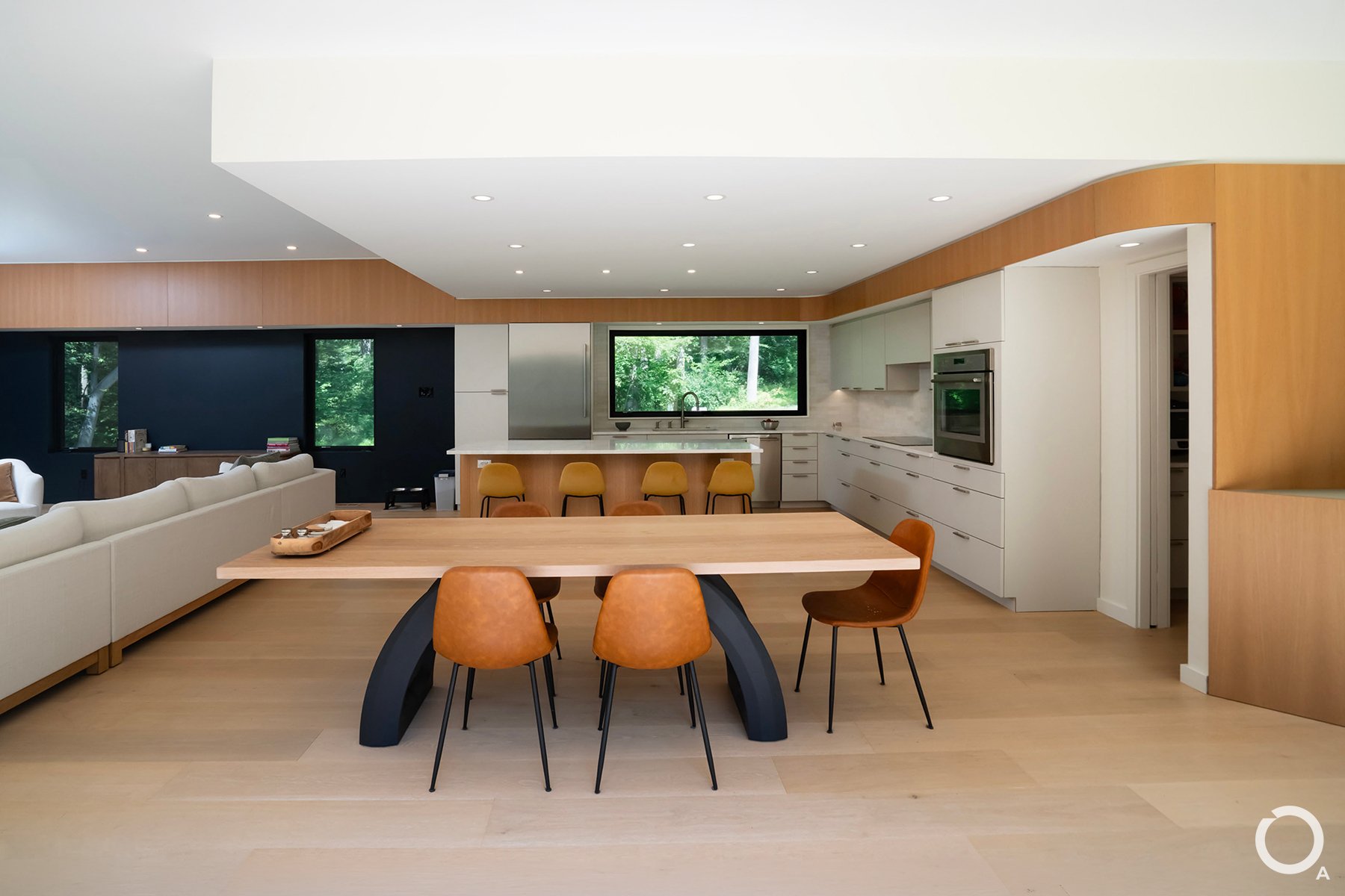 Modern open-plan kitchen and dining room with minimalist white cabinetry, a wood kitchen island, and large windows overlooking the wooded landscape. Photo Credit: Linda McManus Photography
