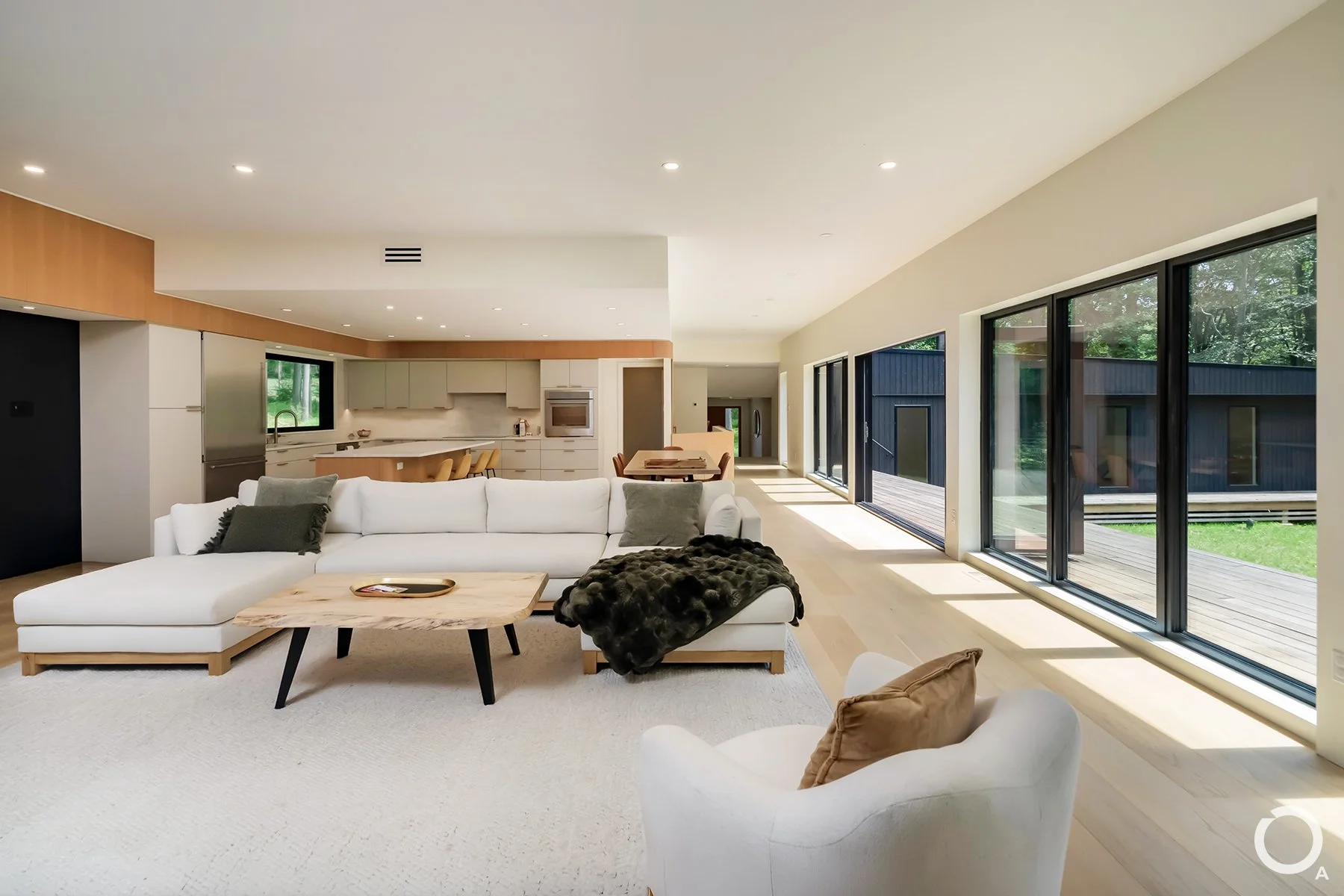 Light-filled modern living room with white sectional seating, floor-to-ceiling glass doors, pale wood flooring, and an open kitchen beyond. Photo Credit: Linda McManus Photography