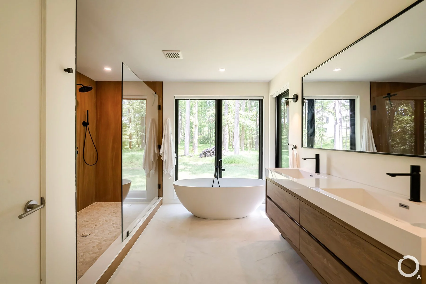 Modern primary bathroom with a freestanding bathtub, double-sink wood vanity, glass-enclosed shower, and large windows overlooking the forest. Photo Credit: Linda McManus Photography