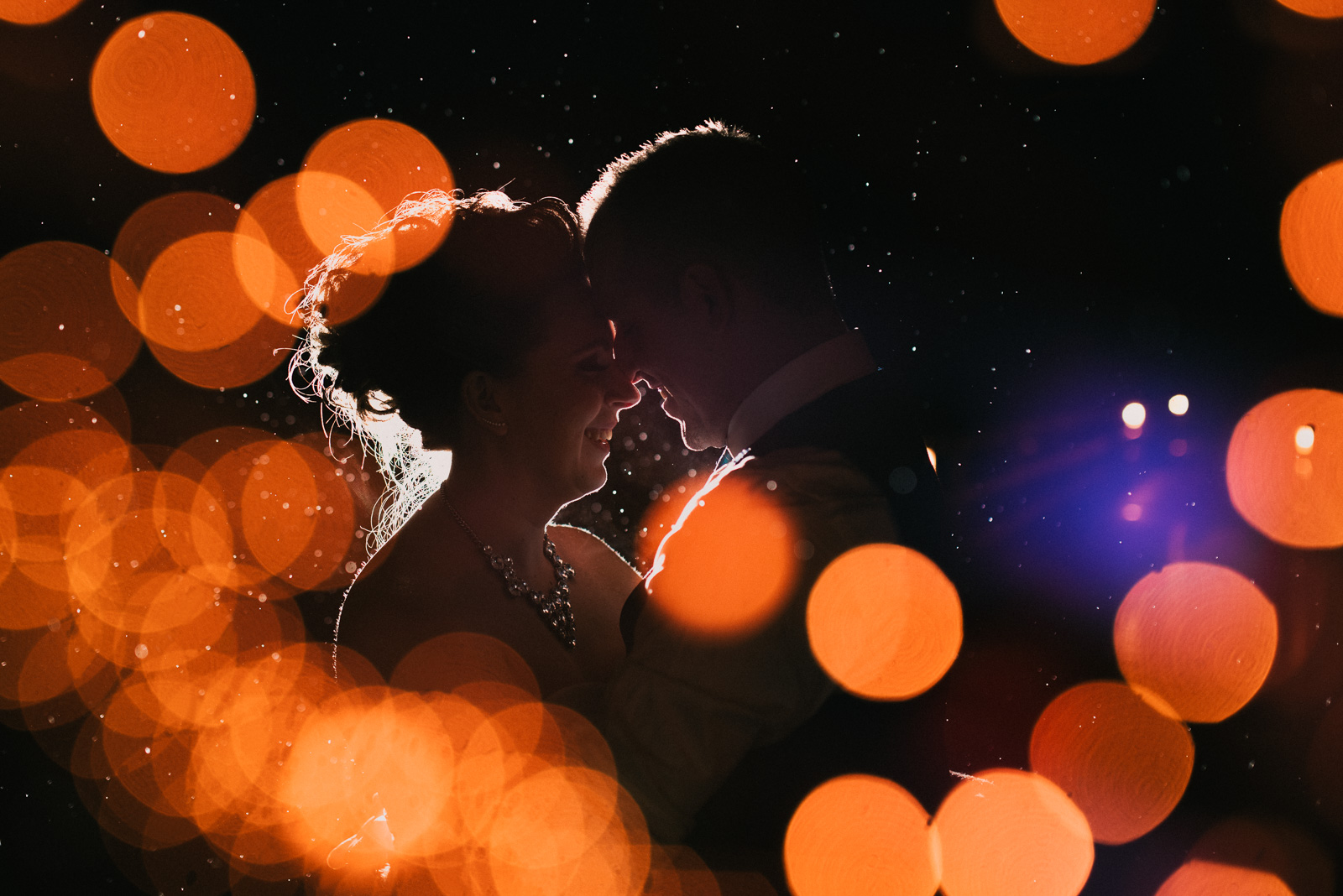 bride and groom standing in the rain at night with blurred string lights at Whonnock Lake Centre in Maple Ridge - Victoria Wedding Photographers