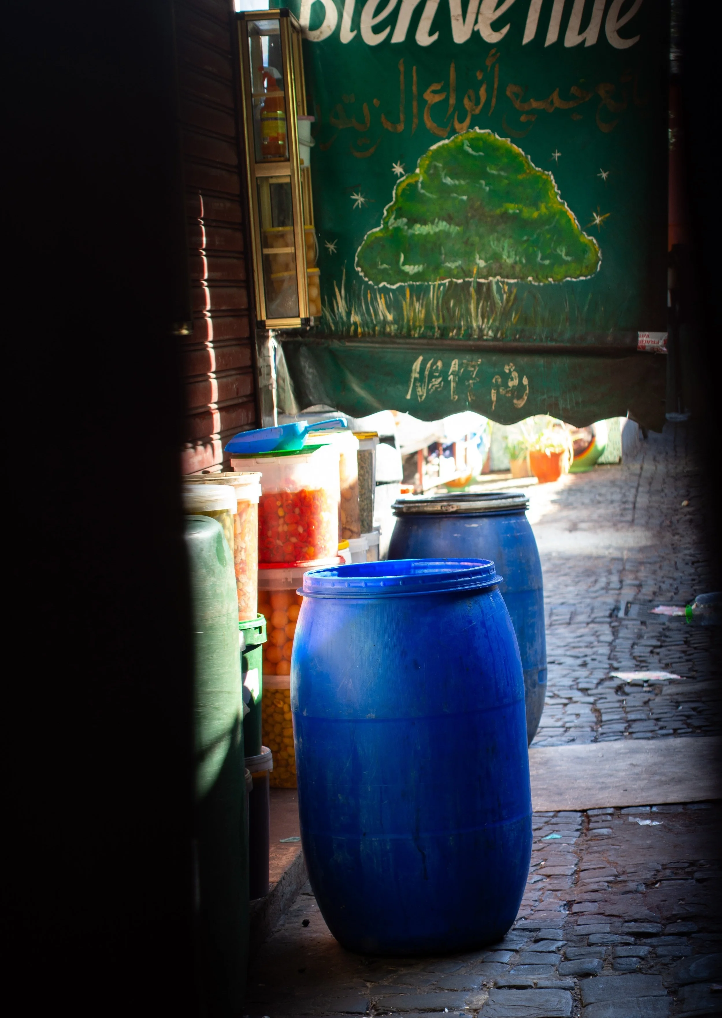 The end of the day; unattended barrels and containers seen through the cracks of the wooden doors of the Great Olive Market, a welcome sign hangs behind them