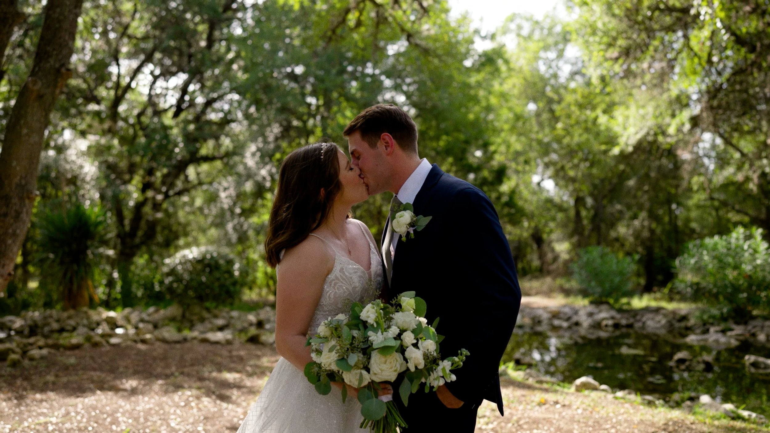 bride and groom kiss with bouquet outside next to trees and pond